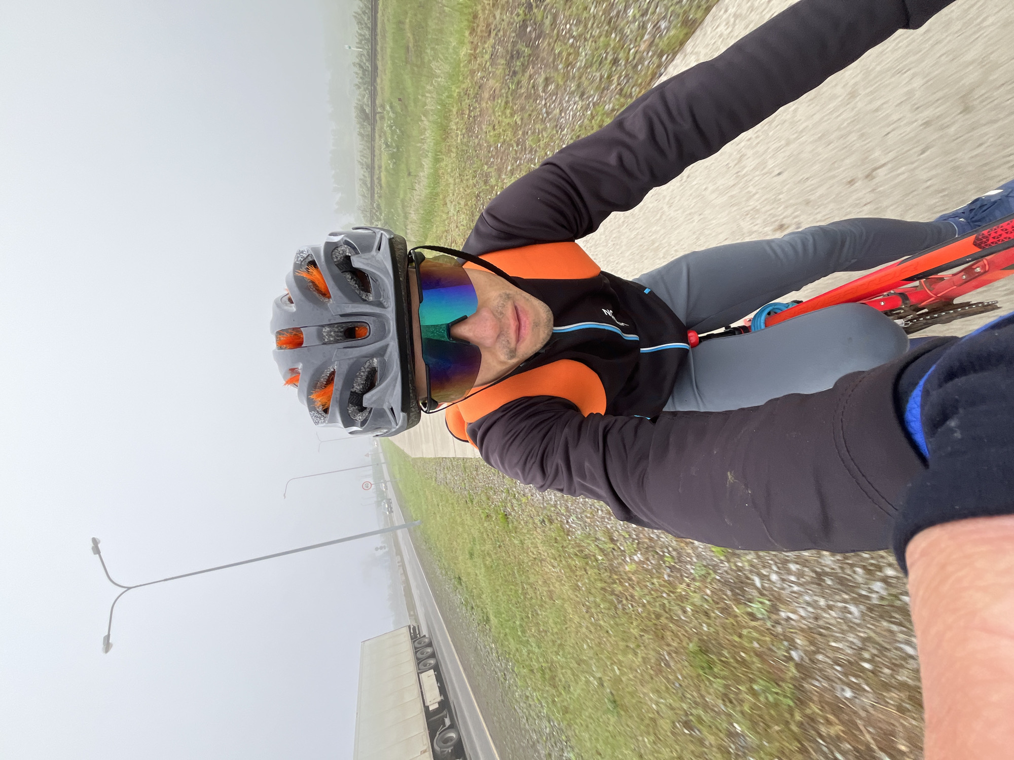 A first-person perspective, rotated 90 degrees, captures a male cyclist riding on a foggy day in Chaikovsky, Russia. The cyclist, likely taking a selfie, wears a grey cycling helmet with bright orange inner padding visible through the vents, and large, reflective, rainbow-tinted sunglasses that obscure his eyes. He is dressed in a black long-sleeved cycling jacket with vibrant orange shoulders and a blue stripe detail across the chest, paired with grey cycling pants. A bright red bicycle frame is visible between his legs, with parts of the chain and possibly a blue seat cushion or bottle holder.
The outdoor scene depicts a paved road to the left and a grassy, gravelly shoulder to the right, suggesting a roadside or highway environment. The weather is characterized by heavy fog or mist, which significantly reduces visibility and creates a soft, muted grey background. In the distance, on the road, a large white truck with a long trailer is partially visible through the haze. A streetlight pole with two luminaires extends from the left side of the frame. A circular traffic sign, potentially indicating a speed limit, is faintly discernible through the fog but unreadable. The overall mood is cool and damp, highlighting the cyclist's brightly colored gear against the monochrome fog.