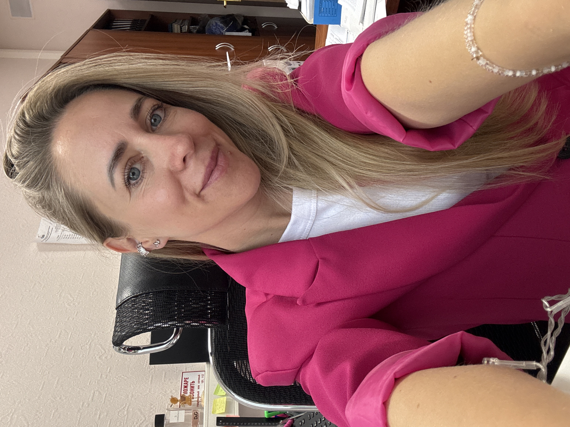 A blonde woman with blue eyes and light skin is pictured in a selfie taken from the chest up. She is wearing a bright pink blazer over a white top and has a delicate bracelet on her right arm. Her hair is long and blonde, falling over her shoulders. She is seated in what appears to be an office environment, with a chair and a desk visible. The background shows office furniture, including a wooden cabinet. A sign with Russian text, possibly related to fire safety ("ПОЖАРЕ БЕЗОПАСНОСТЬ"), is also visible on the wall. The lighting suggests an indoor setting, possibly during the day. The image quality is good, with a slight blur on the hands due to the selfie perspective.