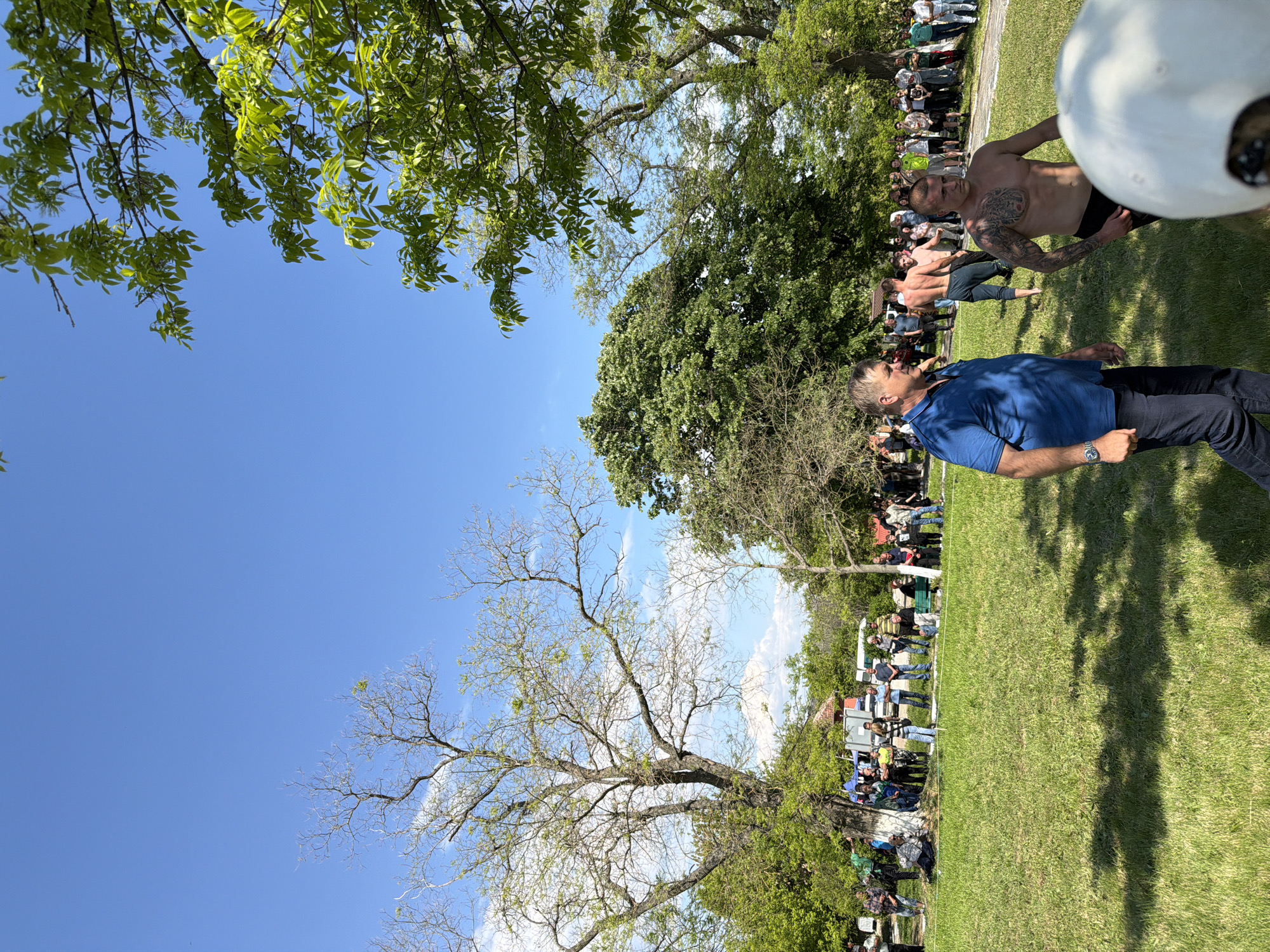 The image depicts an outdoor scene in Dragomir, Bulgaria. A grassy area with scattered trees and a clear blue sky provides the backdrop. A crowd of people stands near what appear to be some temporary structures or food stalls. In the foreground, a man in a blue shirt and dark pants stands facing the crowd. A shirtless man with tattoos on his upper body is positioned near him, partly cut off by the frame, with other shirtless men visible in the background. This suggests some sort of event or competition is taking place. The overall environment appears relaxed and informal.