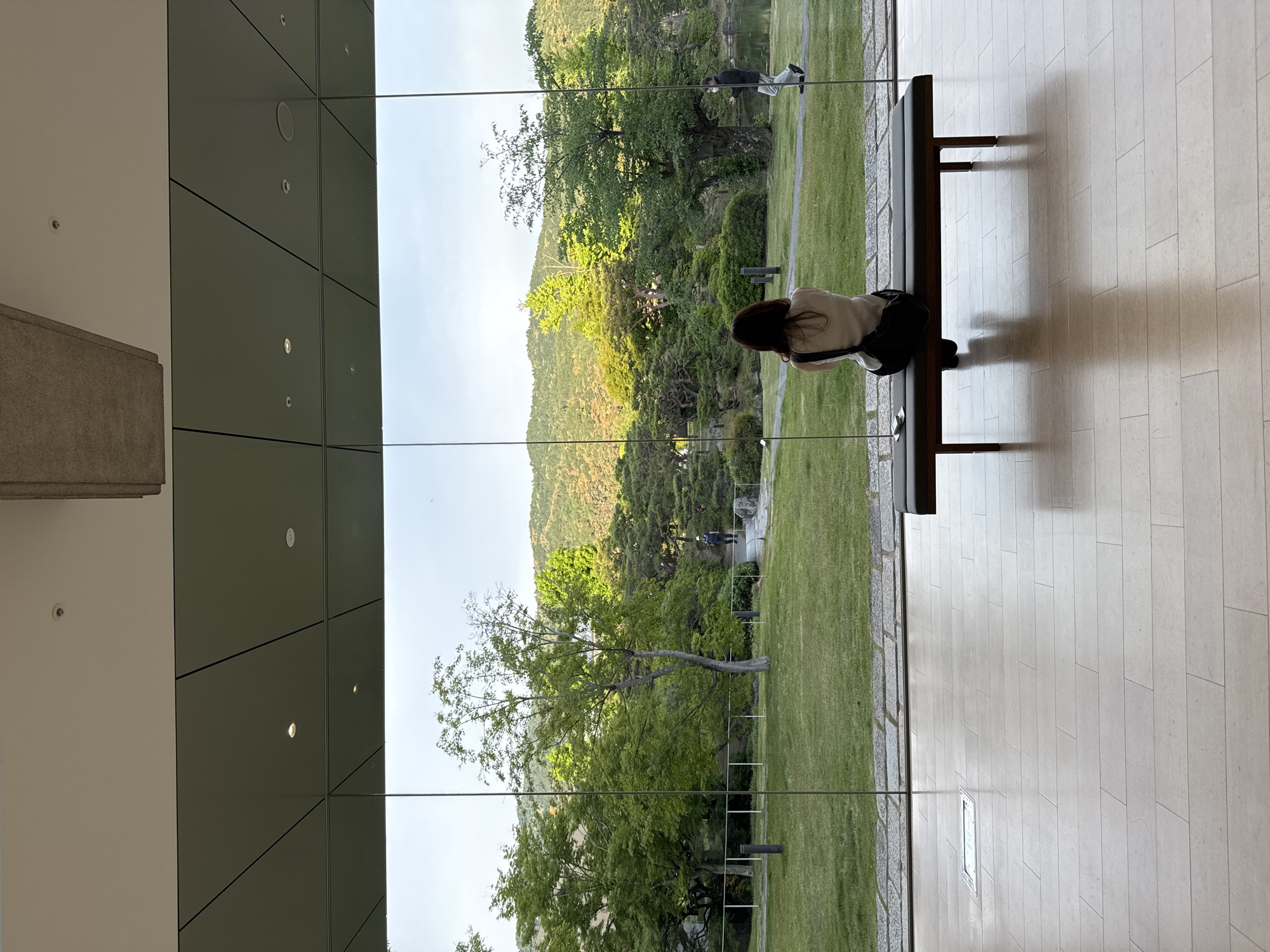 The image shows an interior space in Kyoto, Japan. A woman with long dark hair sits on a bench, seen from the back, facing a large window that reflects a green landscape. The landscape includes a grassy area, trees, and a mountain range. Another person is partially visible in the landscape through the window. The interior has a modern design with tiled floors and paneled walls. The window is composed of multiple panes.