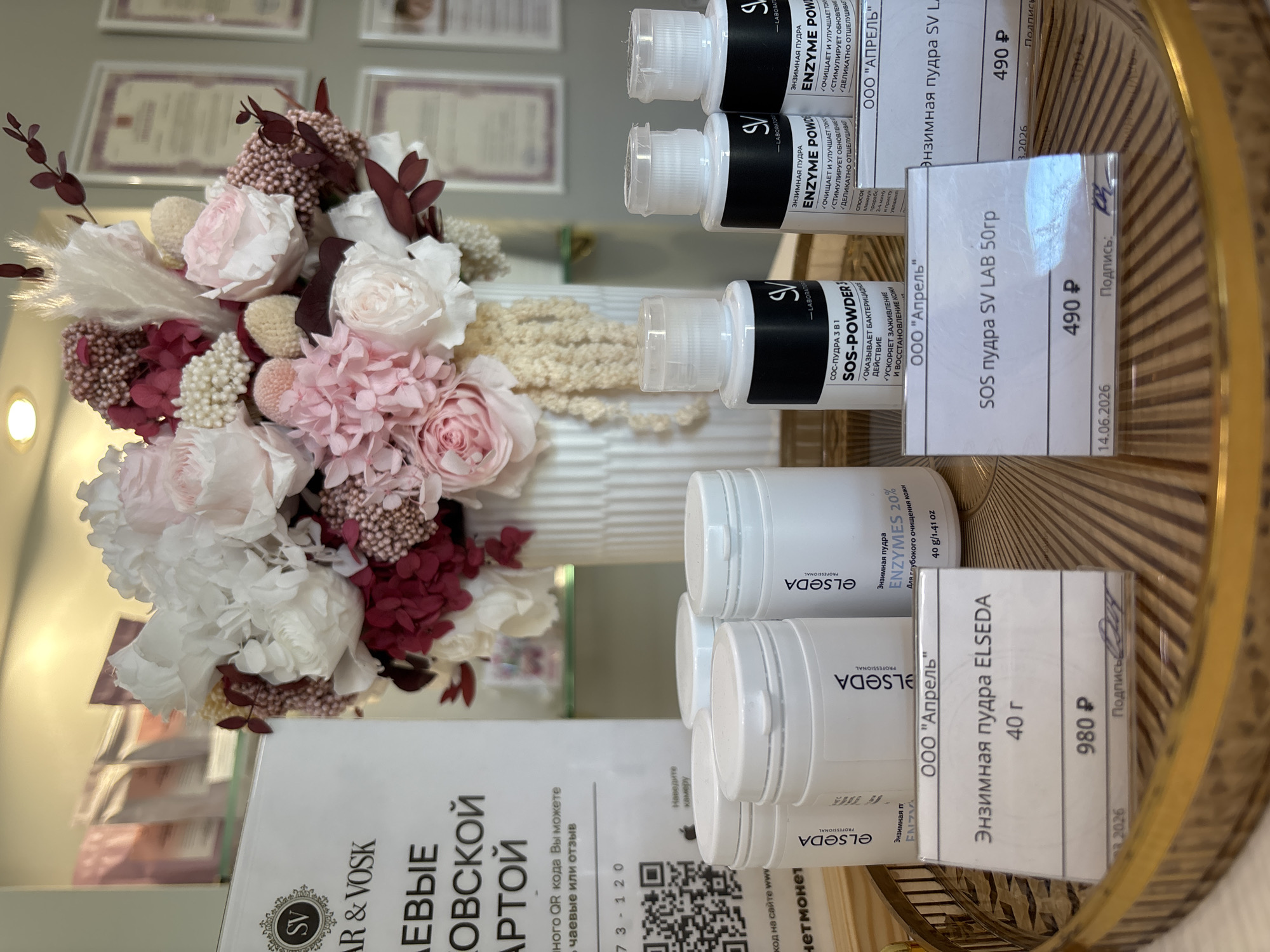 The image shows a close-up of a decorative bouquet of preserved flowers, primarily in shades of pink and white, arranged in a tall, fluted vase. To the right of the flowers, on a reflective golden tray, are several cosmetic products. These appear to be white plastic bottles and jars, some with labels that contain Russian text. One label clearly reads "Энзимная пудра ELSEDA" (Enzyme powder ELSEDA) and mentions a weight of "40 г" (40g). Another product label states "SOS-ПУДРА 3 В1" (SOS-POWDER 3 IN 1) and lists benefits like "ОКАЗЫВАЕТ БАКТЕРИЦИДНОЕ ДЕЙСТВИЕ" (Provides bactericidal effect), "УСКОРЯЕТ ЗАЖИВЛЕНИЕ И ВОССТАНОВЛЕНИЕ КОЖИ" (Accelerates healing and skin restoration). Some of the product labels also include prices like "490 Р" and "980 Р", and dates such as "14.06.2026". There is also a white paper card with similar product information and a handwritten signature. The overall setting suggests a retail or display environment, possibly a beauty salon or a cosmetic store, with a focus on skincare products and floral decor. The lighting is bright, suggesting an indoor setting.