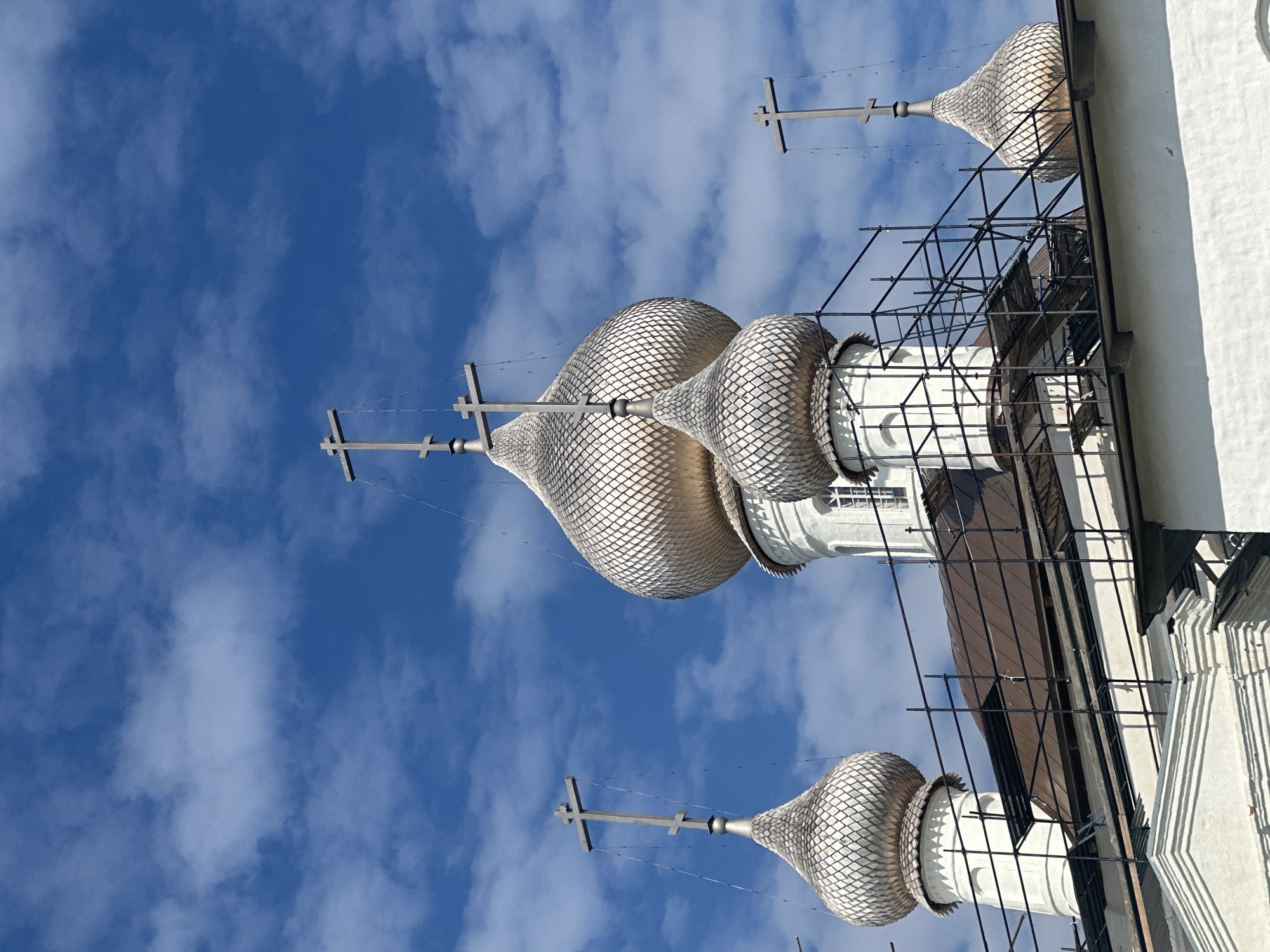 The image shows the roofline of a white building with several onion domes, topped with crosses, against a bright blue sky with scattered white clouds. The domes are covered in a textured, light-colored material, possibly shingles or scales. Scaffolding is visible around some of the domes and the roof. One dome in the center of the image is significantly larger than the others. The building appears to be a religious structure, likely a church, given the presence of domes and crosses. The architecture suggests a Russian Orthodox style. The location is identified as Solovetskiy, Russia.