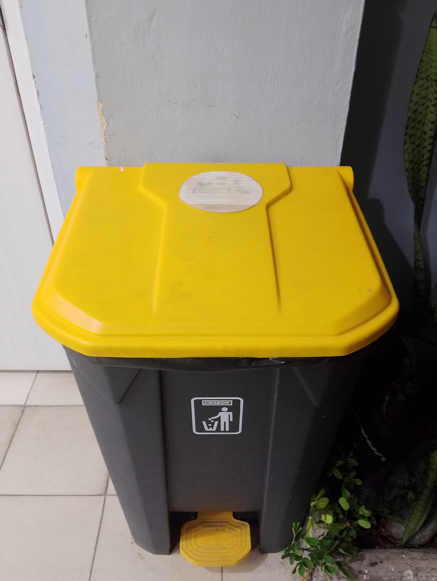A tall, dark gray trash can with a bright yellow lid stands against a light gray wall. The lid has a sticker on it, and the side of the trash can features a white recycling symbol. A yellow pedal is visible at the bottom of the trash can. To the right of the trash can, a snake plant in a pot is partially visible, along with some greenery at the base. The floor is tiled with light-colored squares. The scene appears to be indoors, possibly in an alleyway or service area, and is located in the City of Pematang Siantar, Indonesia.