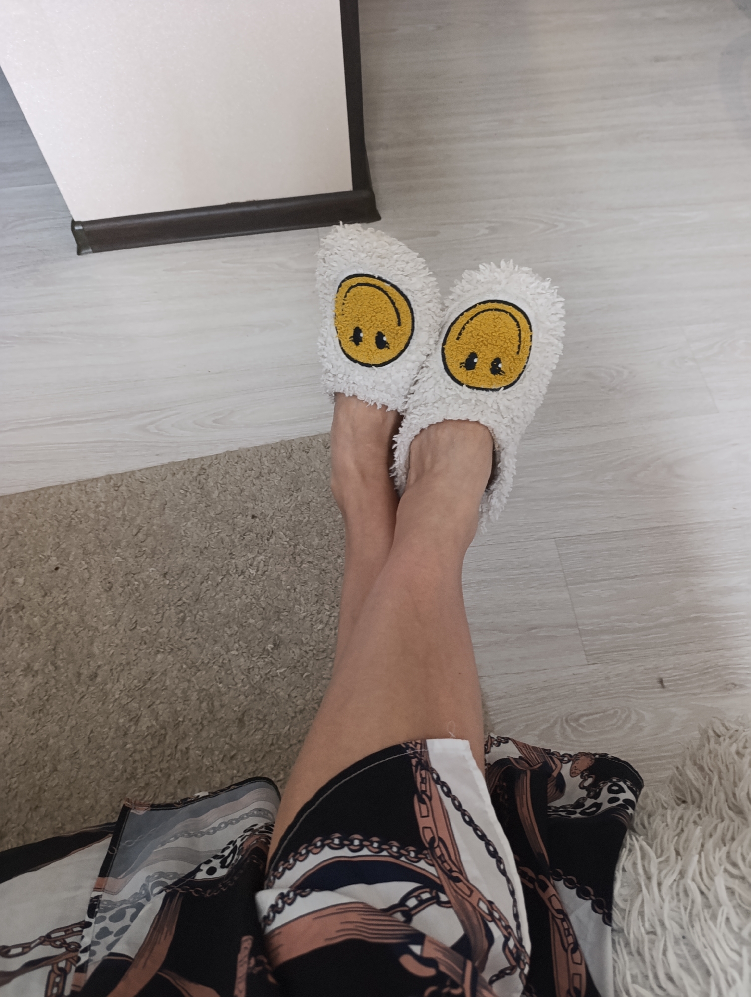 The image shows a person's legs and feet wearing fluffy white slippers with yellow smiley faces. The person is sitting on a carpet next to a light-colored wooden floor. A dark patterned skirt is visible around the person's legs. The background includes a portion of a wall with a dark baseboard. The scene appears to be an indoor residential setting. There is no visible indication of the location being Penza, Russia.