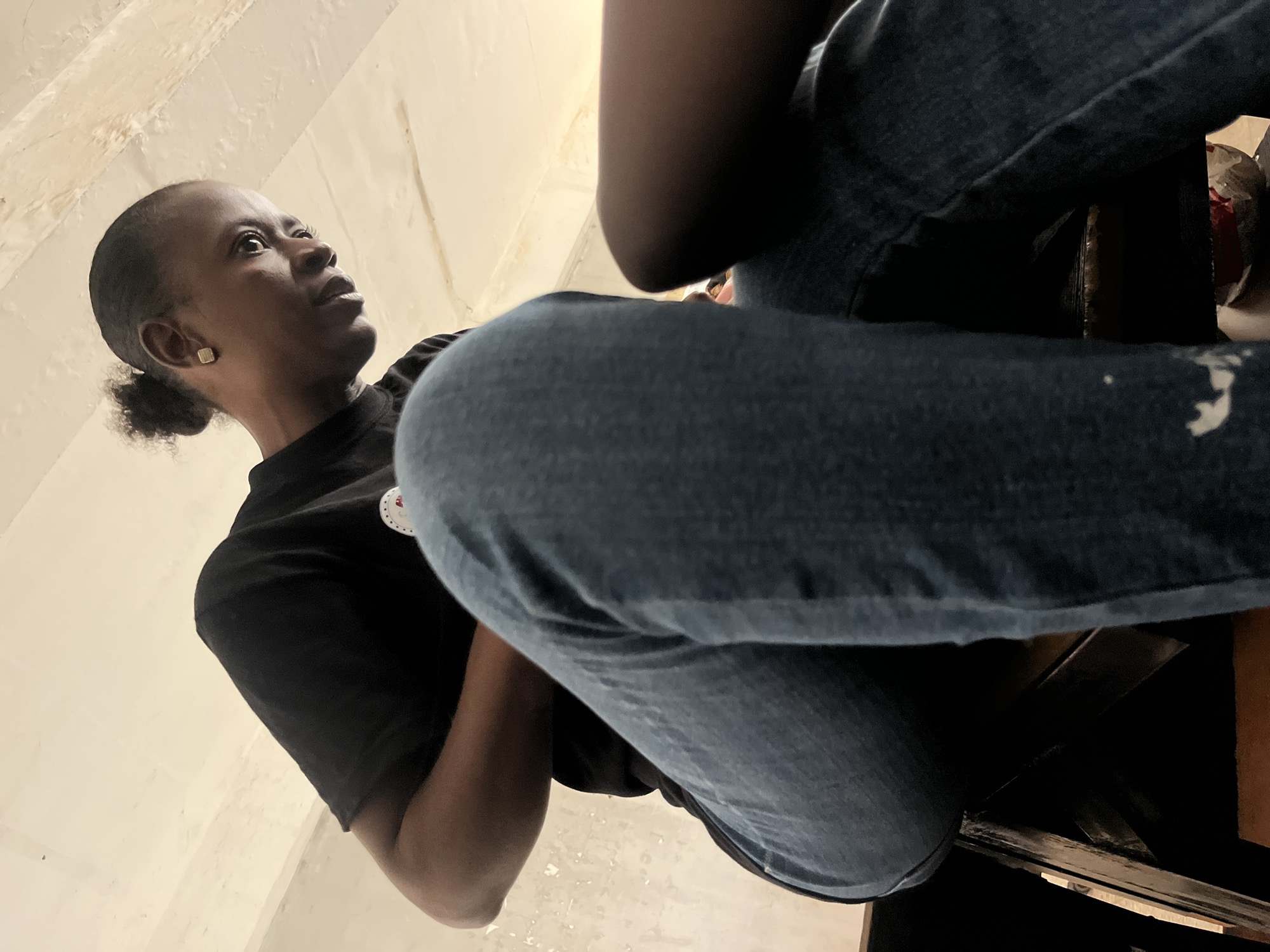 The image is an upward-angled shot of two people, with a focus on the first individual. The first person is a dark-skinned woman with short, dark hair pulled back into a small ponytail. She is wearing a black t-shirt and looking up and to the left. A small stud earring is visible in her left ear. The second person is only partially visible, with their lap and legs prominently featured in the foreground. They are wearing blue jeans. The setting appears to be indoors with off-white walls visible in the background. It is not possible to confirm that the photo was taken in Tudun Nupawa, Nigeria, from the image alone.