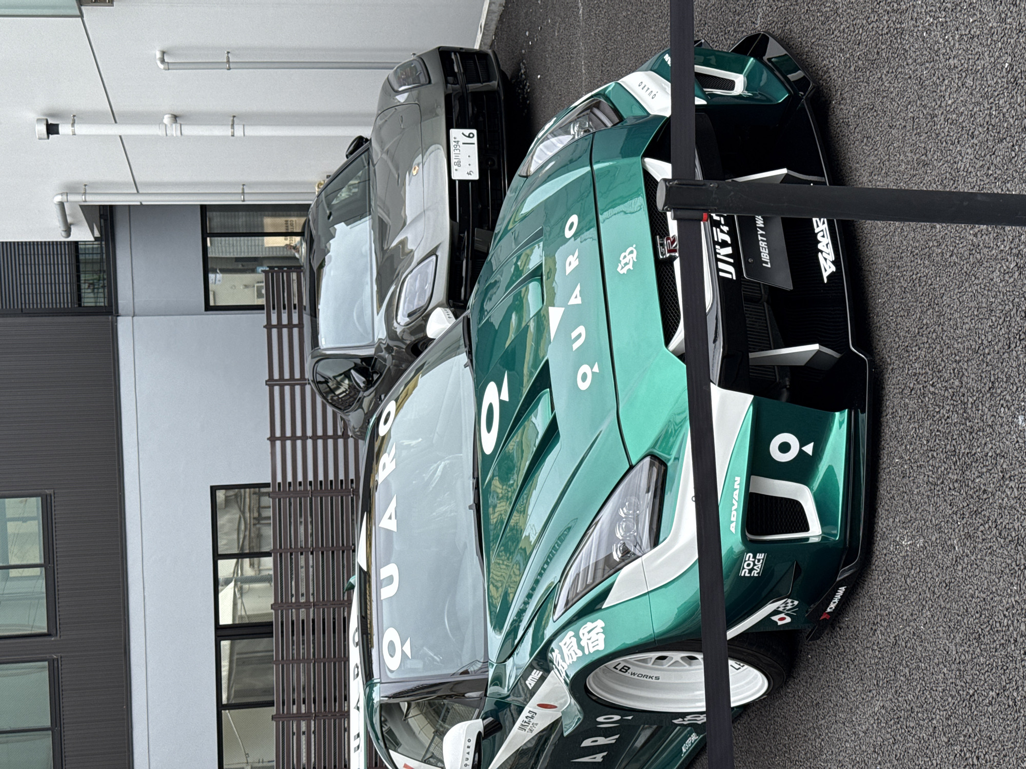 Here's a description of the image:
The image features two cars parked close together on an asphalt surface, possibly a road or parking area. The most prominent car is a green and white Nissan GT-R with distinctive markings, including "QUARO" and "LB WORKS" logos. Adjacent to it is a gray vehicle, partially obscured. A black barrier runs diagonally across the right side of the frame, likely indicating a restricted area or display. The cars are positioned in front of a modern building with gray walls, windows, and metal details. The license plate of the gray vehicle is visible, reading "品川394*ち・・16," which translates to "Shinagawa 394 chi - 16". The presence of a plate registered in Shinagawa suggests the scene is likely located in Japan. The text "原宿" on the green vehicle indicates it is also in Harajuku, located in Shibuya, Japan.
