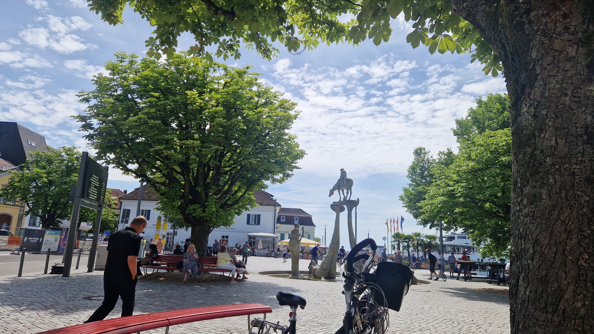 This is a photograph of a public square in Überlingen, Germany, likely near the harbor given the presence of a large white boat in the background. The scene is set on a sunny day with scattered white clouds in a blue sky. Large trees with lush green foliage are prominent in the foreground and midground, with one particularly large tree trunk on the right side of the frame.
In the midground, a statue featuring a person on horseback atop a pedestal is visible. To the left of the statue, a building with white walls and blue shutters stands. A group of people are seated on a red bench in front of this building, appearing to be resting or socializing. A man in a black shirt and pants is walking away from the viewer towards the right side of the frame, his hands in his pockets.
In the foreground, a red bench is positioned partially in view, and a bicycle with a black basket is parked. The ground is paved with light-colored cobblestones. Further in the background, there are more trees, flags, and a harbor with a boat. Other people are scattered throughout the square, some appearing to be walking or standing. A large digital display board is visible on the left side of the image. The overall atmosphere appears to be relaxed and public.