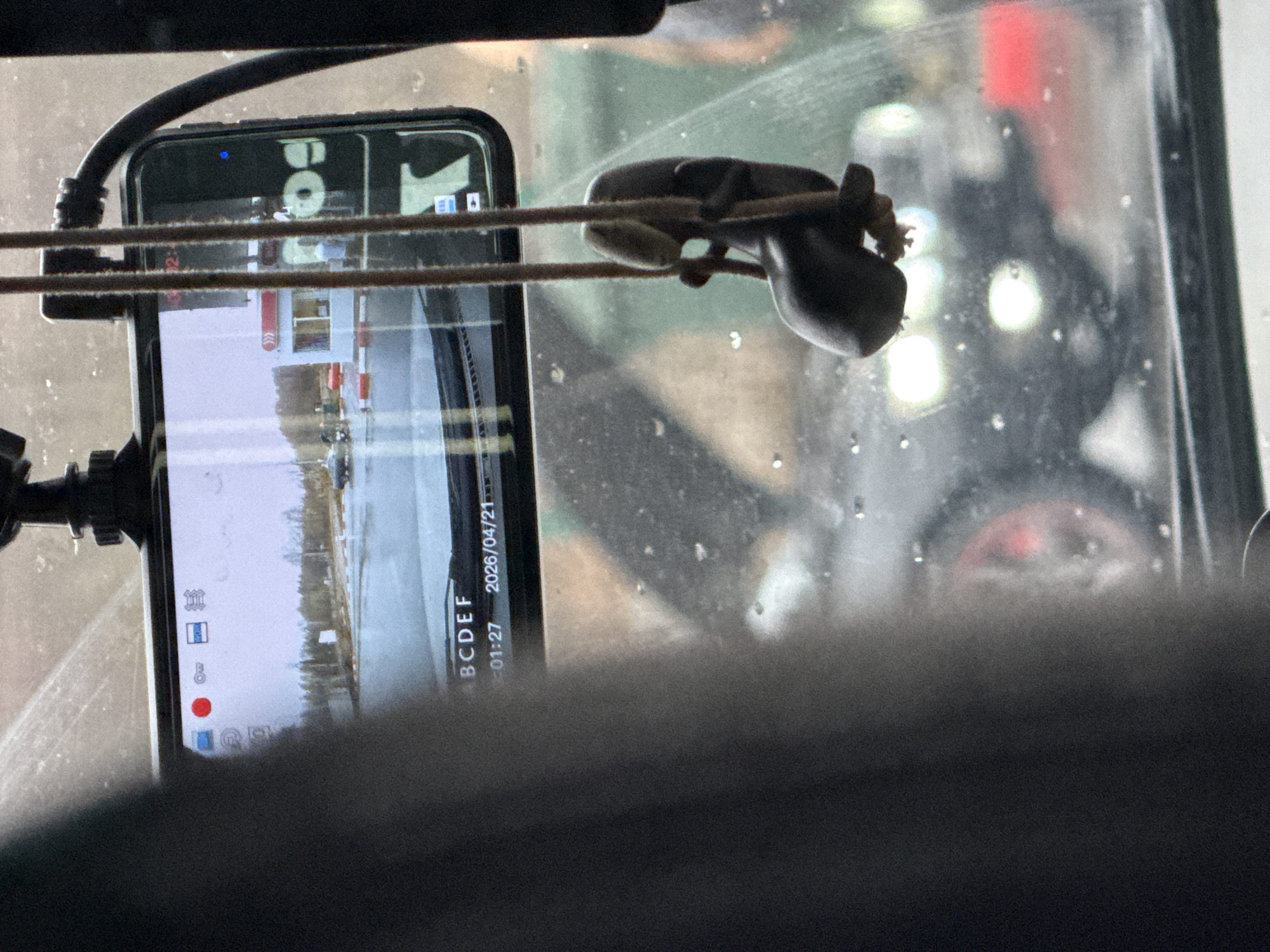 The image depicts a view from inside a vehicle, looking outwards through a dusty or moisture-speckled windshield. In the foreground, a black smartphone is mounted vertically, actively recording video. A red recording dot is visible on its screen, along with several icons typical of a camera interface. The phone screen displays the date "2026/04/21" and a timestamp "01:27", likely indicating the video duration rather than the time of day, given the daylight outside. The text "BCDEF" is also visible at the bottom right of the screen, and "Bol" (possibly part of "Bolt" or a similar word) at the top.
A small, dark, stylized object resembling a frog or toad figurine is suspended by a string or wire in front of the windshield, positioned between the smartphone and the glass. Two taut, parallel wires or strings run horizontally across the frame, passing in front of both the phone and the figurine.
The phone's screen reflects the external landscape, revealing a scene with bare trees, possibly snow or ice on the ground, and some distant structures featuring red and white striped elements. This landscape suggests a rural or semi-rural setting in Klenovskoe, Russia, consistent with early spring conditions where remnants of winter might still be present. The light appears diffused, possibly indicating an overcast day. Parts of the vehicle's dark interior frame the top and bottom of the shot.