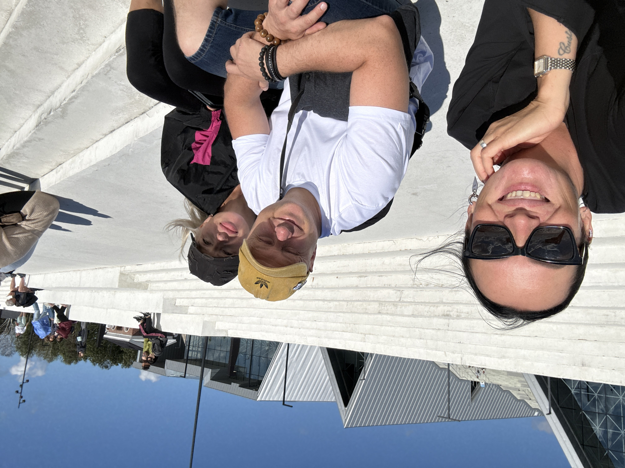 The image depicts a group of people in an outdoor setting, appearing to be at a modern architectural complex with a large, stepped, white concrete structure. The background features buildings with distinctive angular, silver-colored roofs and glass facades. The sky is clear and blue. In the foreground, three individuals are prominently featured, seated on the concrete steps.

On the left, a woman with dark hair pulled back, wearing sunglasses and a black top, is smiling and resting her chin on her hand. She is wearing a wristwatch and has visible tattoos on her arm. To her right are a man and a woman. The man, wearing a yellow baseball cap and a white t-shirt, is looking towards the camera with a slight smile. He has several bracelets on his wrist and is wearing denim shorts. Next to him, a woman with blonde hair, wearing a black jacket with pink accents and a black cap, is also seated. The three in the foreground appear to be posing for a selfie.

In the mid-ground and background, other individuals are seen sitting and walking on the steps and around the complex, suggesting it's a public space. The overall scene suggests a sunny day at a contemporary urban or cultural site. The location is identified as Kronstadt, Russia.