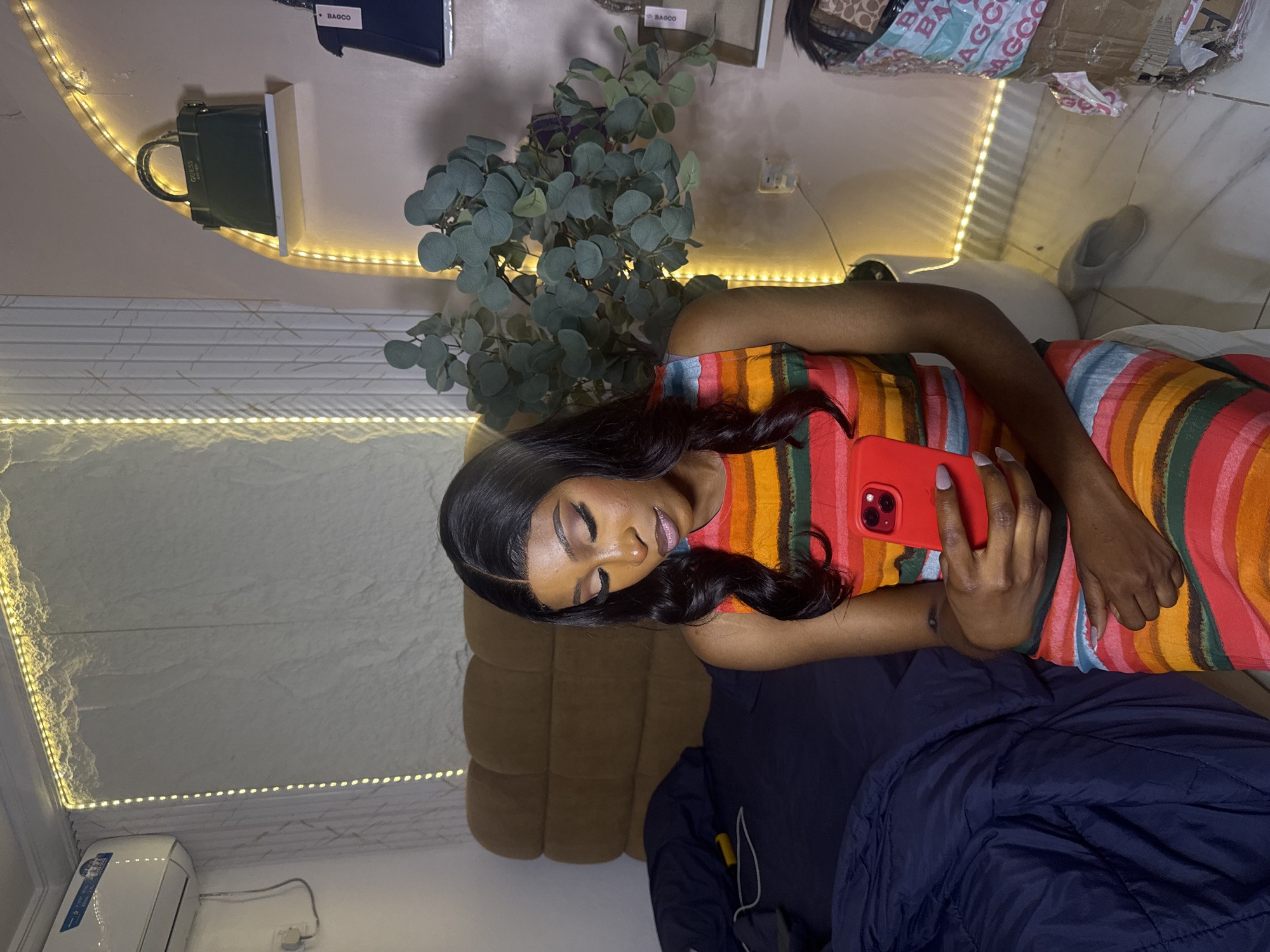 The image depicts a medium shot of a person with dark skin and long, dark hair, sitting on a bed and holding a red smartphone. The person is wearing a sleeveless dress with horizontal stripes in red, orange, yellow, green, and blue. Their makeup includes dramatic eye shadow and eyeliner. The background shows a bedroom with a large, tufted brown headboard, a dark blue duvet on the bed, and an air conditioning unit on the wall. Decorative lighting, in the form of LED strips, illuminates the wall behind the bed and an arched shelving unit. On the shelf, there are two handbags, one green and one dark blue. A large artificial eucalyptus plant is positioned to the right of the person. The floor is tiled, and a white planter is visible. The scene appears to be indoors in what could be a residential setting in Abuja, Nigeria.