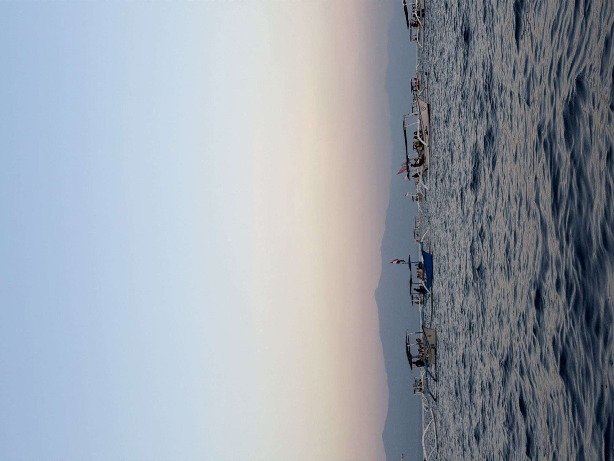 The image depicts a serene seascape at dusk, featuring a line of traditional Balinese outrigger boats sailing on calm waters. The boats, some carrying passengers, are silhouetted against the backdrop of a pastel-hued sky transitioning from pale blue to soft pink and orange. In the distance, the faint outlines of mountains are visible, suggesting a coastal setting. The water is a deep blue, with gentle ripples reflecting the muted light. The scene evokes a sense of tranquility and the end of a day in a tropical paradise, likely a popular tourist destination in Indonesia, such as the waters off of Bali or Lombok.
