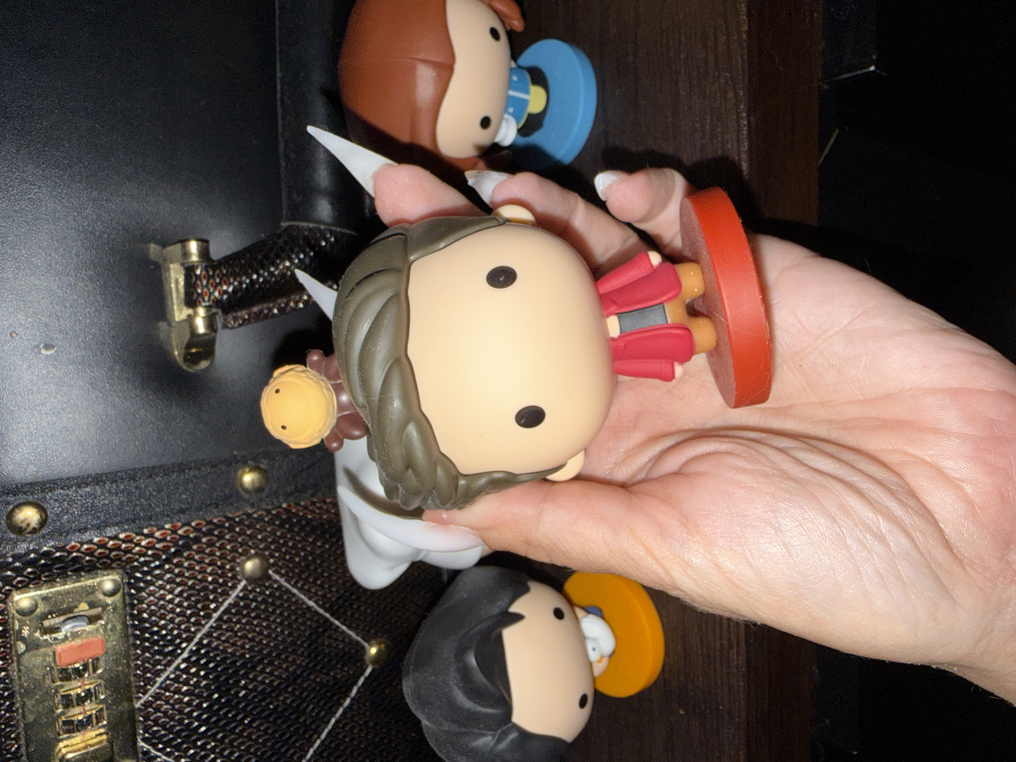 A hand is holding a collection of collectible figures in front of a dark, textured background with brass accents. The figures appear to be of cartoonish characters. One figure, held prominently in the center, has brown hair, a red jacket over a tan shirt, and is standing on a red circular base. To the left of this figure, another figure with black hair and a white shirt is visible, standing on an orange circular base. To the right, a third figure with brown hair and a blue dress is present, standing on a blue circular base. A small, yellow, spherical creature with brown limbs is perched on top of the central figure's head. The background appears to be a piece of furniture with metallic embellishments. The scene is indoors, with the lighting casting shadows that suggest an overhead light source. The image does not contain any information indicating a location in Odesa, Ukraine.