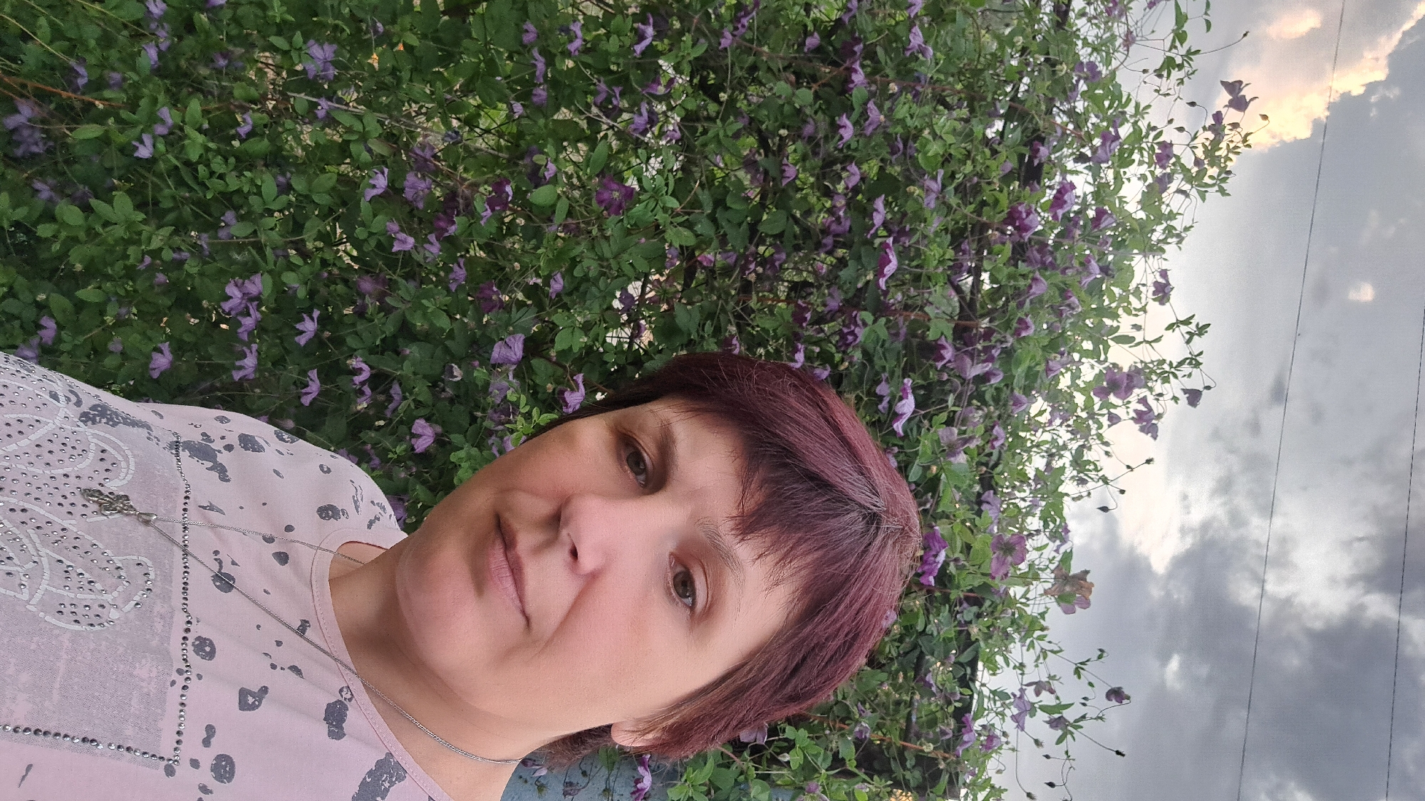 A woman with short, dark red hair and fair skin is taking a selfie in front of a large bush with many small purple flowers. She is wearing a light pink t-shirt with a grey, abstract design. A silver necklace with a pendant is visible around her neck. The background shows a grey, cloudy sky with some lighter patches of sky. The image appears to be taken outdoors, possibly in a garden or park. The location is in the city of Chernetsk, Russia.