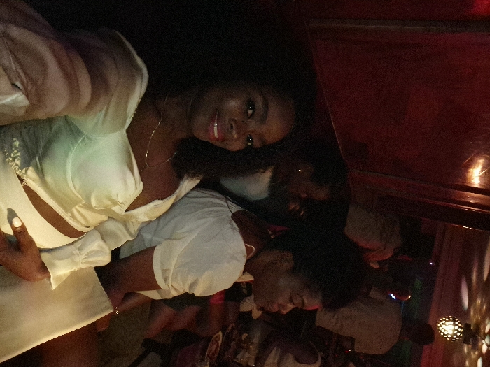 Two smiling women pose in a dimly lit bar or restaurant. The first woman, closest to the viewer, wears a light-colored satin top with cutouts. Her hair is dark and curly, and she wears hoop earrings. The other woman wears a white button-down shirt. Other people are seen in the background, along with glasses and bottles on the bar. A red wall is visible on the right, and a mirrored disco ball is partially visible in the bottom right corner.