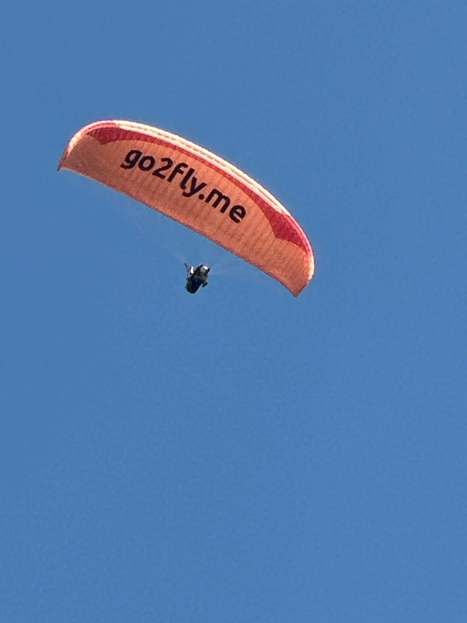 A paraglider is captured in mid-flight against a clear blue sky. The paraglider's canopy is orange with a red trim and features the text "go2fly.me" in black. A single paraglider pilot is suspended beneath the canopy, appearing as a dark silhouette. The image is a photograph taken outdoors, likely during daylight. The specific location in Boreti, Montenegro, is not discernible from the visual information provided in the image.