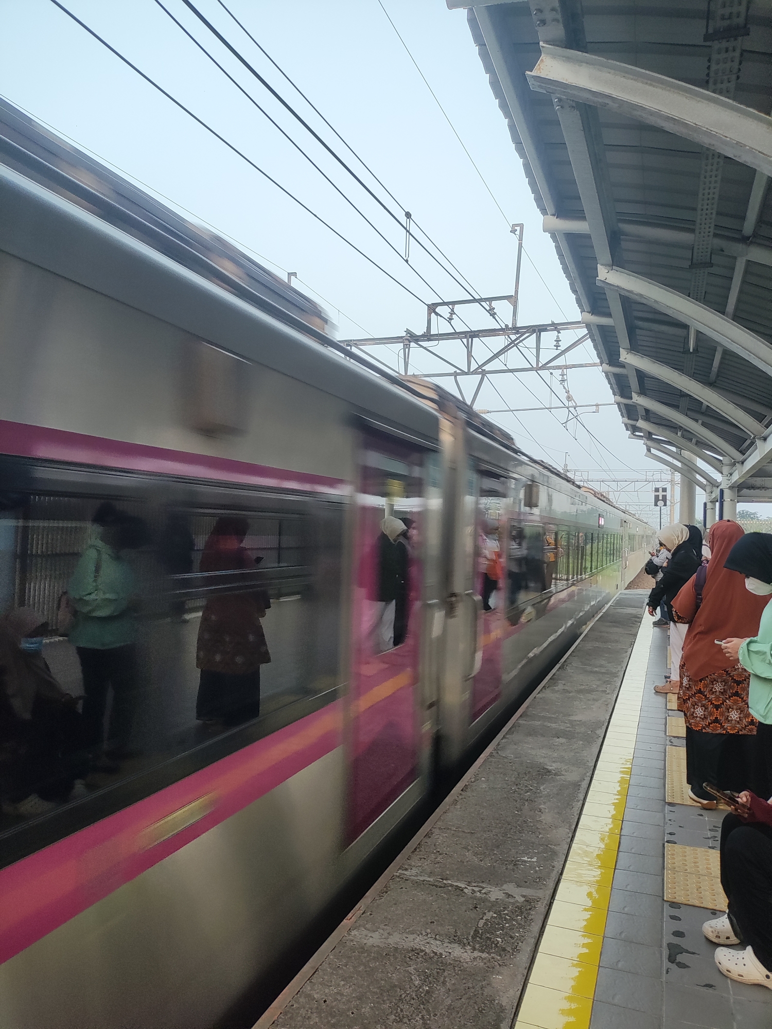 The image captures a train station platform in Telagamurni, Indonesia, with a train moving past. A train, depicted with motion blur, is seen on the tracks. The platform is paved with tiles, featuring a yellow safety line near the edge. Several people are visible on the platform, some standing and waiting, while others are seated. They are dressed in casual clothing, with some wearing headscarves. The environment appears overcast, with a covered waiting area supported by metal structures. Overhead, power lines for the train are visible against a pale sky. The scene is a snapshot of everyday life at a public transportation hub.
