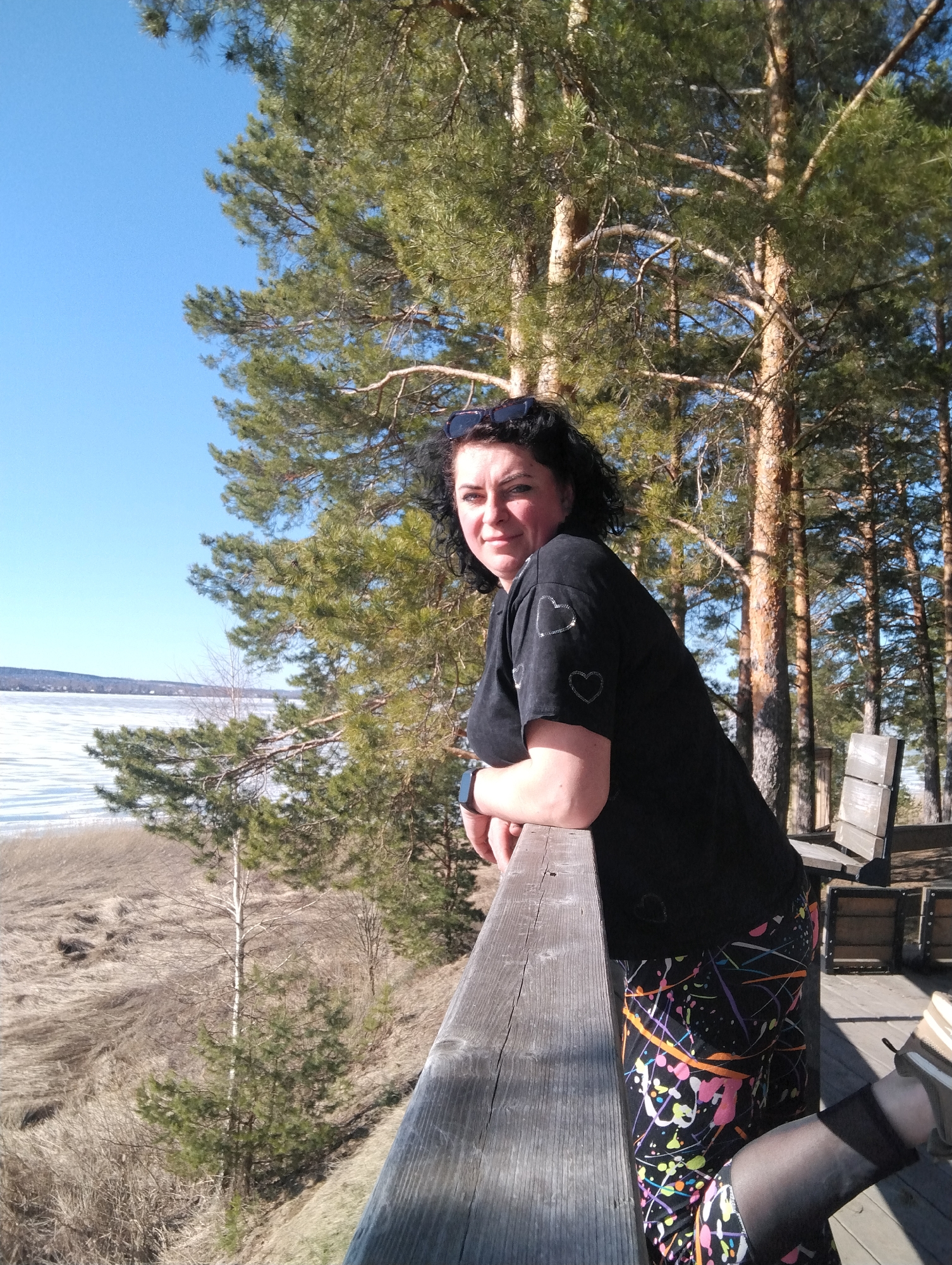 A woman stands on a wooden railing on a sunny day in Votkinsk, Russia, overlooking a partially frozen lake. She wears a black T-shirt with heart embellishments, leggings with colorful paint splatters, and sheer black socks with a tan shoe. Sunglasses are perched on her head. In the background are pine trees and a wooden bench. Below the railing is a grassy slope with some bare trees and shrubs. The sky is blue.