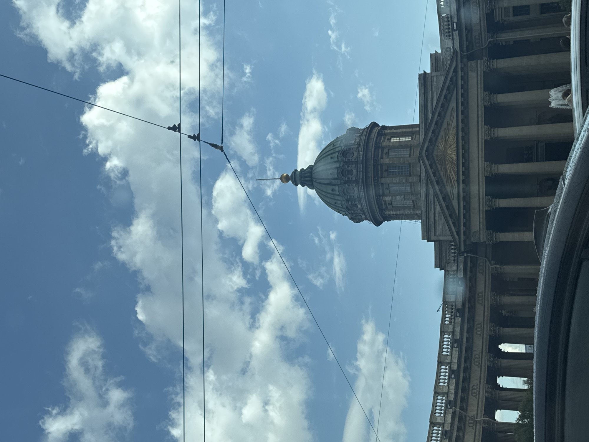 The image shows the Kazan Cathedral in Saint Petersburg, Russia, on a cloudy day. The cathedral's dome and colonnade are visible, along with overhead wires. In the foreground, the roof of a car is visible, partially obscuring the lower part of the cathedral's facade. A fountain is visible to the right of the image, and a few people are standing near it. The sky is blue with scattered white clouds. The overall impression is one of a historic landmark viewed from street level.