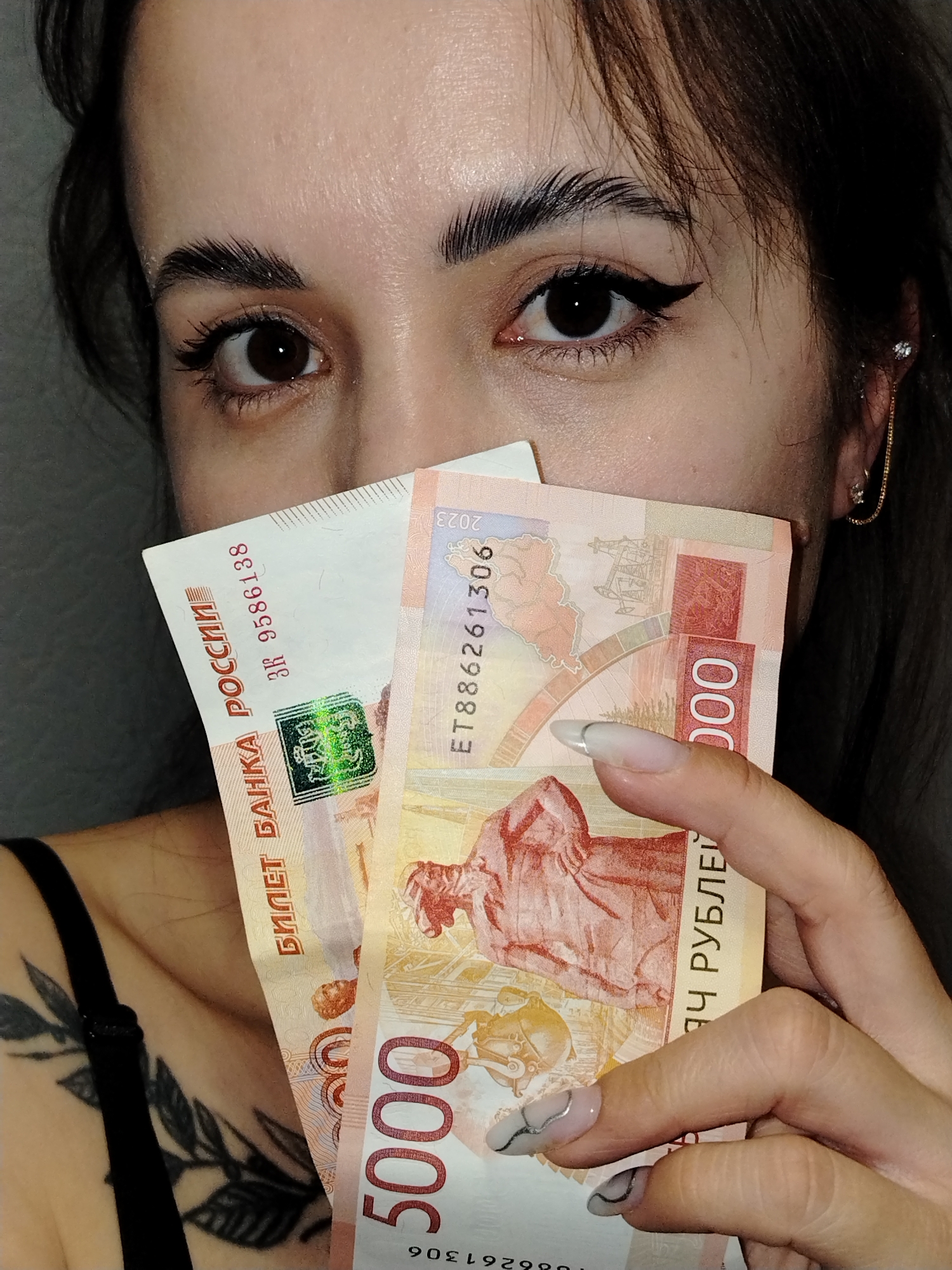 A person is holding two Russian Ruble banknotes in front of their face. The banknotes are of the 5000 denomination. The person has dark hair, visible eye makeup, and a tattoo on their shoulder. Part of their face, including their eyes and eyebrows, is visible above the banknotes. The location mentioned, Noviy, Russia, is relevant to the currency shown in the image.