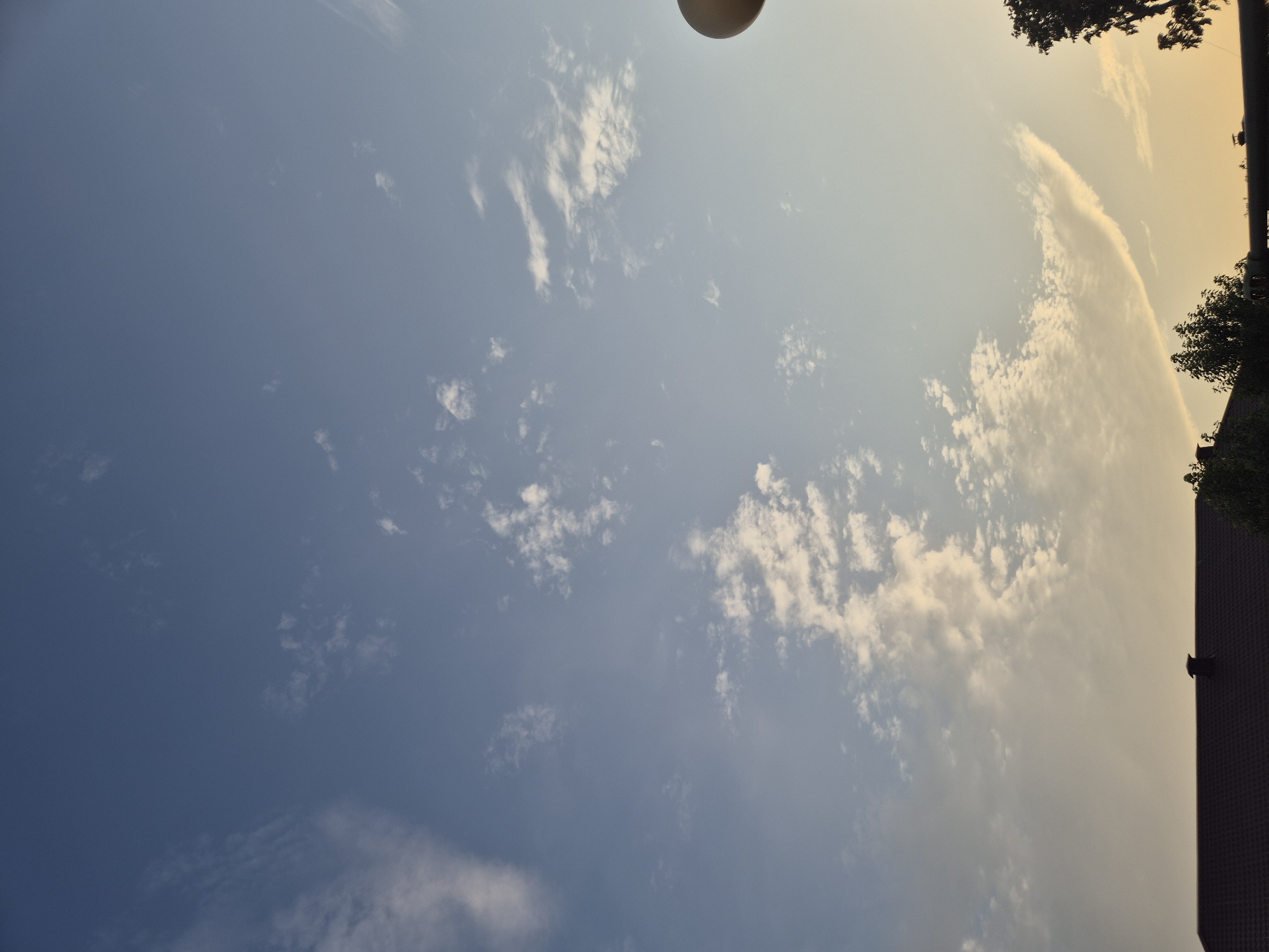 The image shows a clear sky with scattered white clouds, transitioning to a pale orange hue near the horizon, suggesting it might be near dawn or dusk. The lower portion of the image features the dark silhouette of rooftops, trees, and a chimney. A portion of a light fixture is visible on the far right side of the frame. The setting appears to be a residential area within the Парыцкі сельскі Савет in Belarus. There are no people or visible activity depicted in the scene.