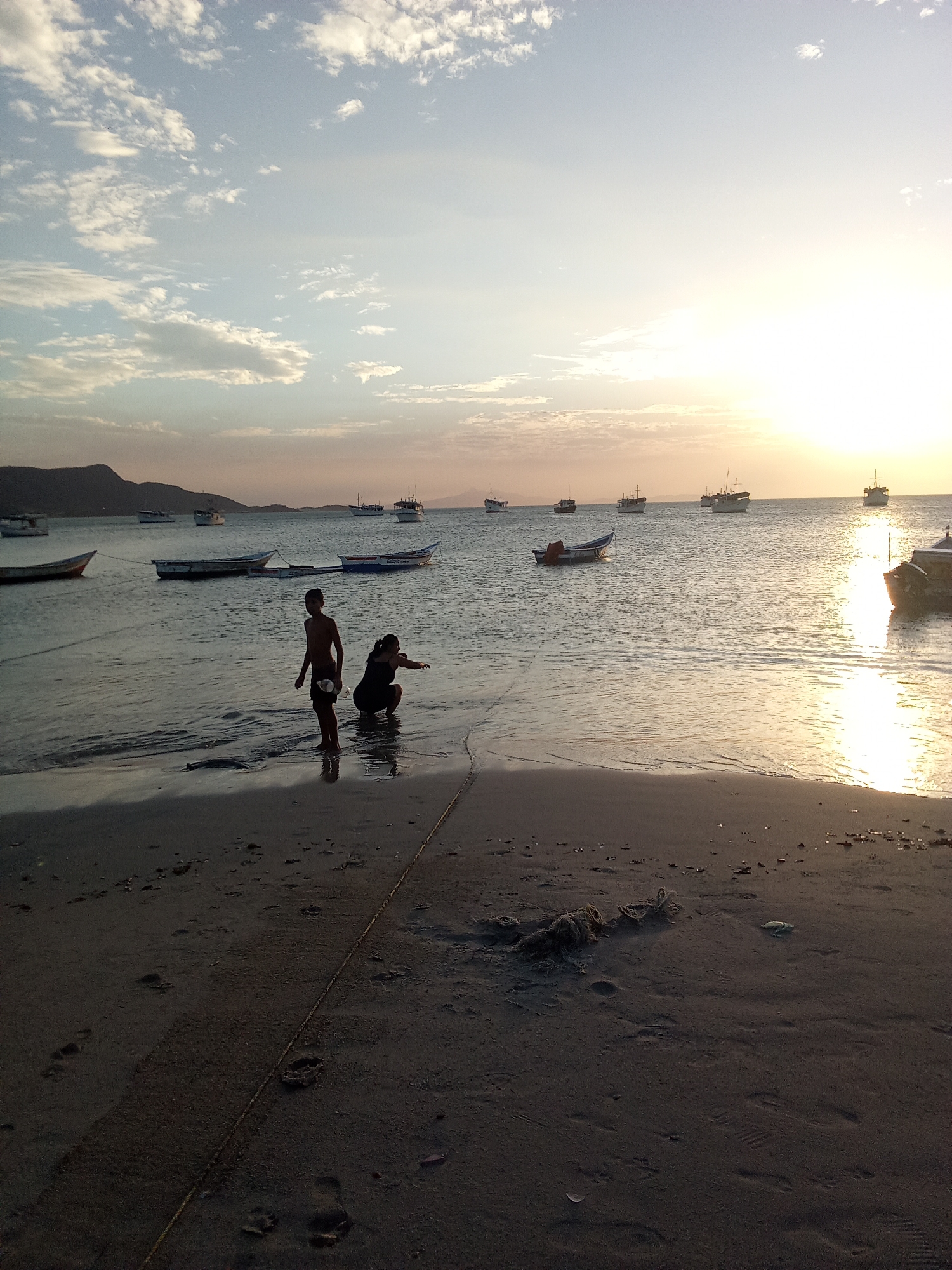 The image shows a beach scene at sunset in Juan Griego, Venezuela. The sky is filled with light clouds and a bright setting sun. Several boats are anchored in the calm water, with the silhouettes of distant hills visible. On the beach, two people, likely a child and an adult, are wading in the shallow water. A rope stretches across the sand towards the water's edge. The foreground of the beach is dark, showing footprints and some debris.