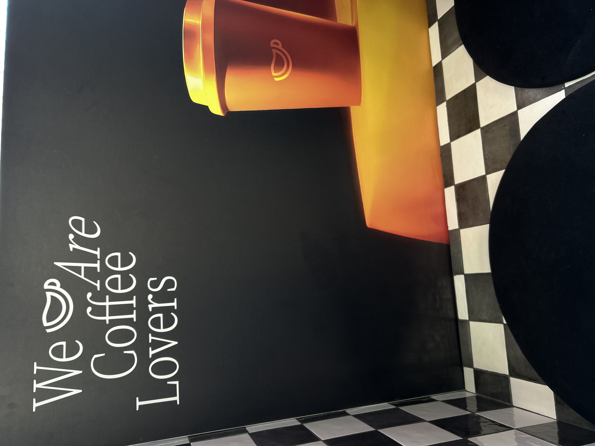 The image depicts a corner of a room, likely indoors. On the left is a black wall with the white text "We Are Coffee Lovers" accompanied by a coffee cup graphic. Above the text, there is an orange and yellow stylized image of a coffee cup. The floor and adjacent wall are covered in a black and white checkered pattern. To the right, partially obscuring the checkered wall, are two large, round black objects, possibly seating or decorative elements. The image appears to be taken in a commercial or retail setting. There is no indication that this image is in Saint Petersburg, Russia.