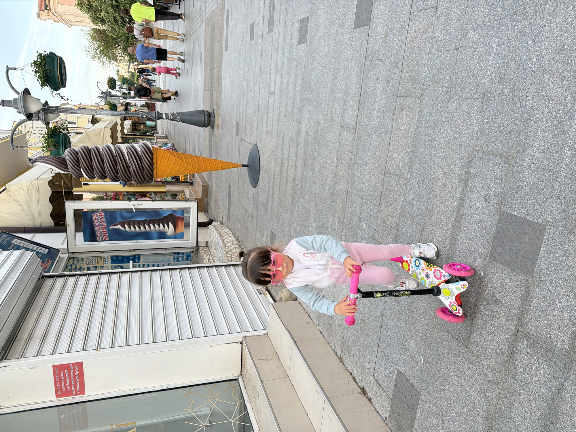 A young girl with sunglasses is standing on a floral-patterned scooter on a stone-paved street. The girl is wearing pink pants and a light-colored jacket. To her left, a closed store with a sign in Bulgarian is visible, along with a large, decorative ice cream cone display. In the background, people are walking along the street. The sign on the store indicates it is "The Dubai Perfumery," and the store is located on "Raina Knyaginya" street number 14, near the museum in Burgas, Bulgaria.