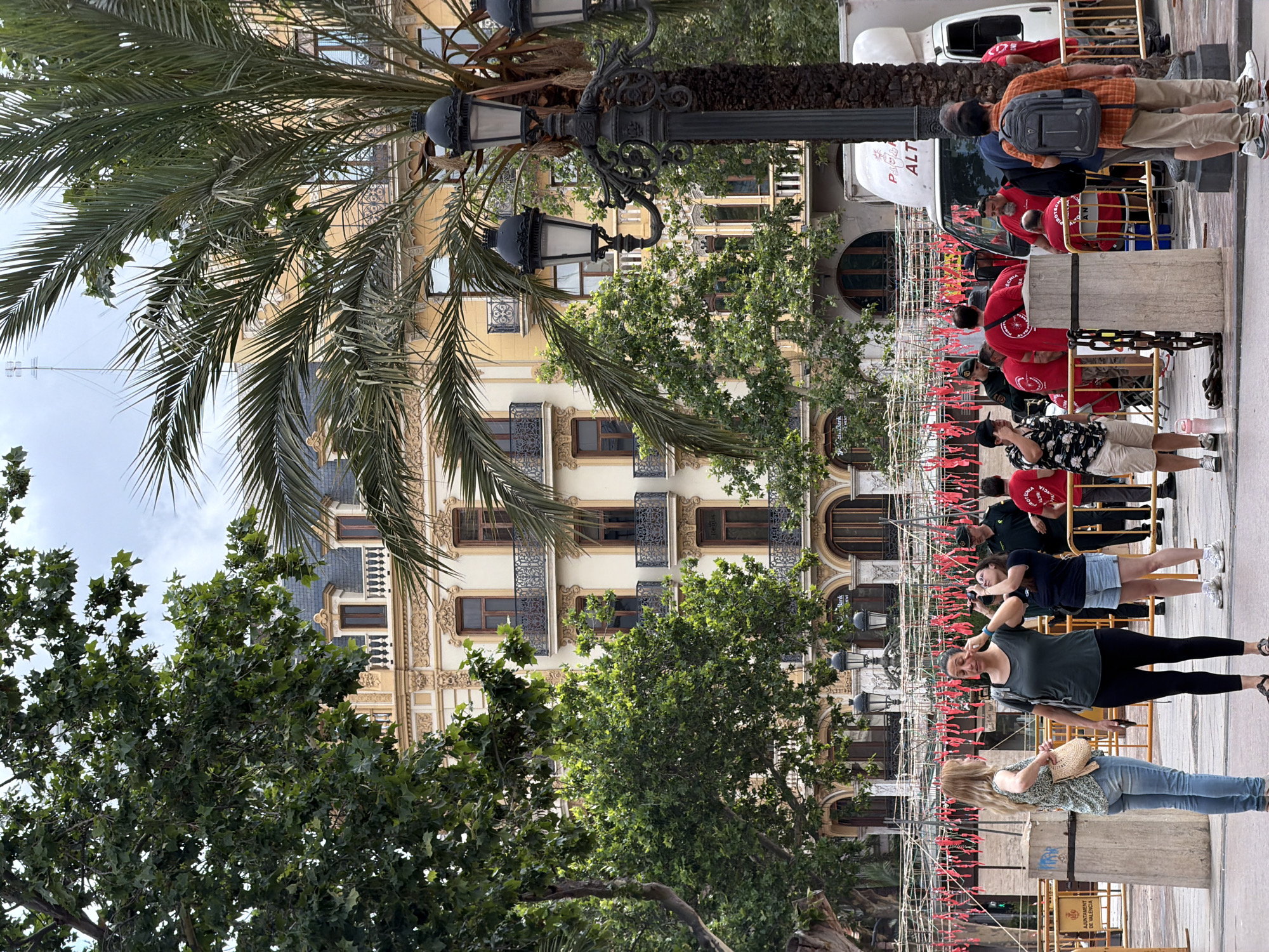This detailed image captures a daytime scene in the Plaza del Ayuntamiento (Town Hall Square) in València, Spain, during preparations for a pyrotechnic event. In the background, a grand, multi-story historical building with elaborate Beaux-Arts architecture dominates the scene. This is the former Banco de València, now a BBVA branch. Its facade is light cream-colored with intricate gold-toned decorative relief work, dark-framed windows, and numerous wrought-iron balconies. The upper section features a dark grey mansard roof with dormer windows. Two large trees frame the view: a dense green deciduous tree on the left and a tall palm tree on the right, partially obscuring a multi-lamp black ornate street light. In the mid-ground, a complex structure of white poles and taut wires is erected, from which numerous small red cylindrical objects, identified as pyrotechnic elements, hang in rows. This is characteristic of a 'mascletà' setup, a traditional percussive fireworks display in València. A white panel van, partially visible behind the setup and the palm tree, bears the legible text "PIROTECNIA ALT," confirming its association with fireworks. In the foreground and mid-ground, several individuals are present: * On the left, a woman with light-colored hair, wearing a patterned short-sleeved top and blue jeans, stands holding a light-colored woven bag, engaged in conversation. * Beside her, another woman with dark hair, dressed in a dark green t-shirt and black leggings, adjusts what appears to be an earplug in her right ear, a common practice before loud pyrotechnic events. She has a dark backpack. * Behind them and to the right, a third woman with dark hair, wearing a dark top and denim shorts, holds a black camera pointed towards the fireworks setup, potentially filming or photographing. * Further right, a man in a dark green uniform, possibly security or local police, with a cap and a badge on his sleeve, observes the scene. * A group of men in red t-shirts, some with indistinct text, are gathered near the fireworks setup and the white van. These individuals appear to be event staff or pyrotechnicians. One man in a patterned short-sleeved shirt and light shorts stands slightly apart, adjusting his dark baseball cap. * On the far right, a man with his back to the camera, wearing an orange plaid shirt, khaki pants, and a grey backpack, stands near the palm tree. * The ground is paved, and various metal barriers and concrete bollards