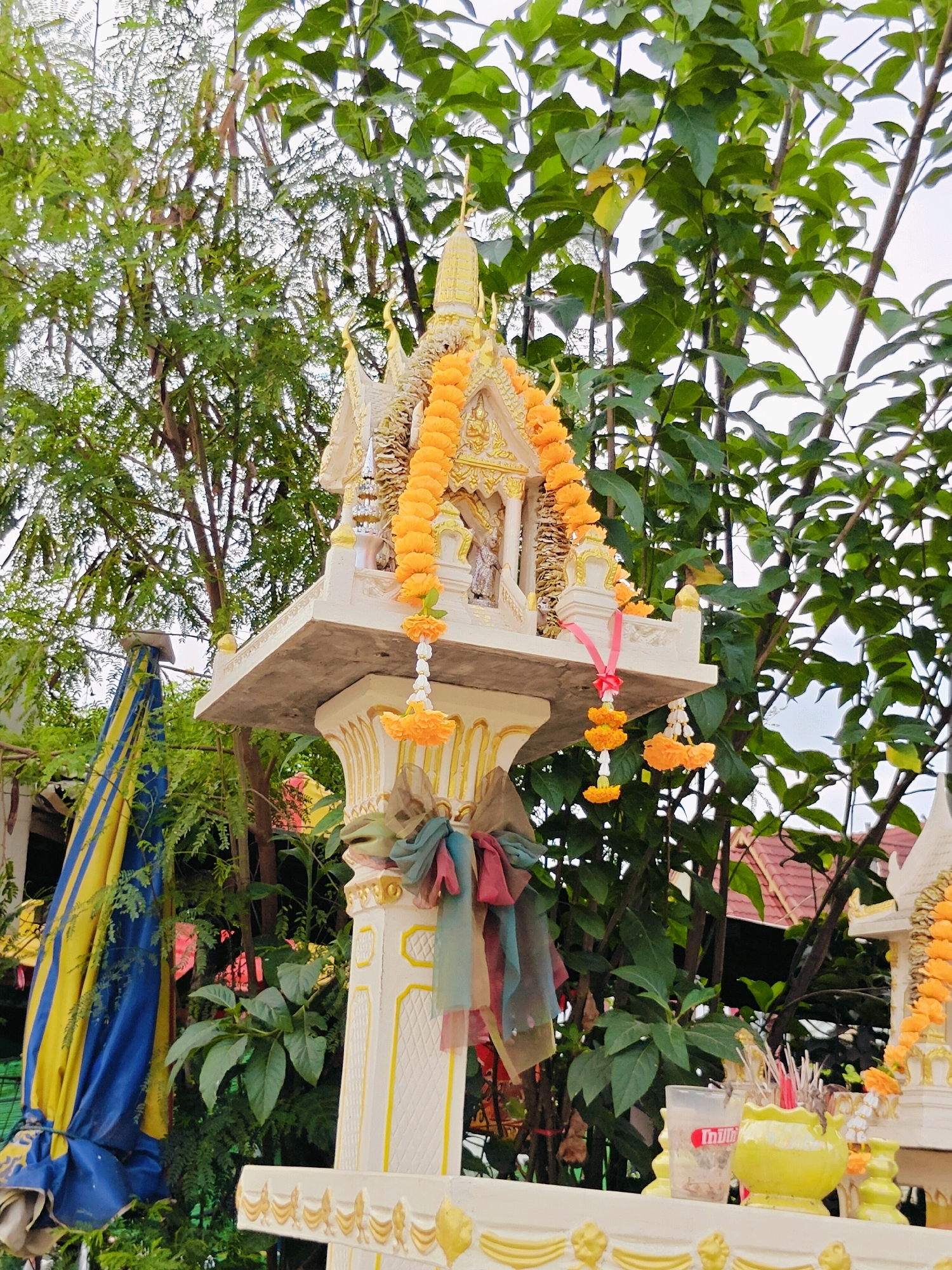 A Thai spirit house, elaborately decorated with garlands of marigolds and fabric streamers, stands prominently outdoors. The spirit house is white with gold accents and topped with a tiered, pagoda-like structure. It is adorned with orange flower garlands that drape over its sides. Red and pink ribbon is tied to one of the garlands. Several colorful fabric strips in shades of green, blue, red, and brown are tied to the pillar supporting the main structure. To the left, a large, folded blue and yellow striped umbrella or fabric is leaning against a dark surface. The spirit house is situated in a lush environment with abundant green foliage and trees, suggesting an outdoor setting in a tropical climate. In the background, a red-tiled roof of a building is visible. On the platform in front of the spirit house, there are various offerings, including a clear plastic cup with a red label, a yellow incense burner with several red-tipped incense sticks, and other small decorative items. The sky above is overcast.