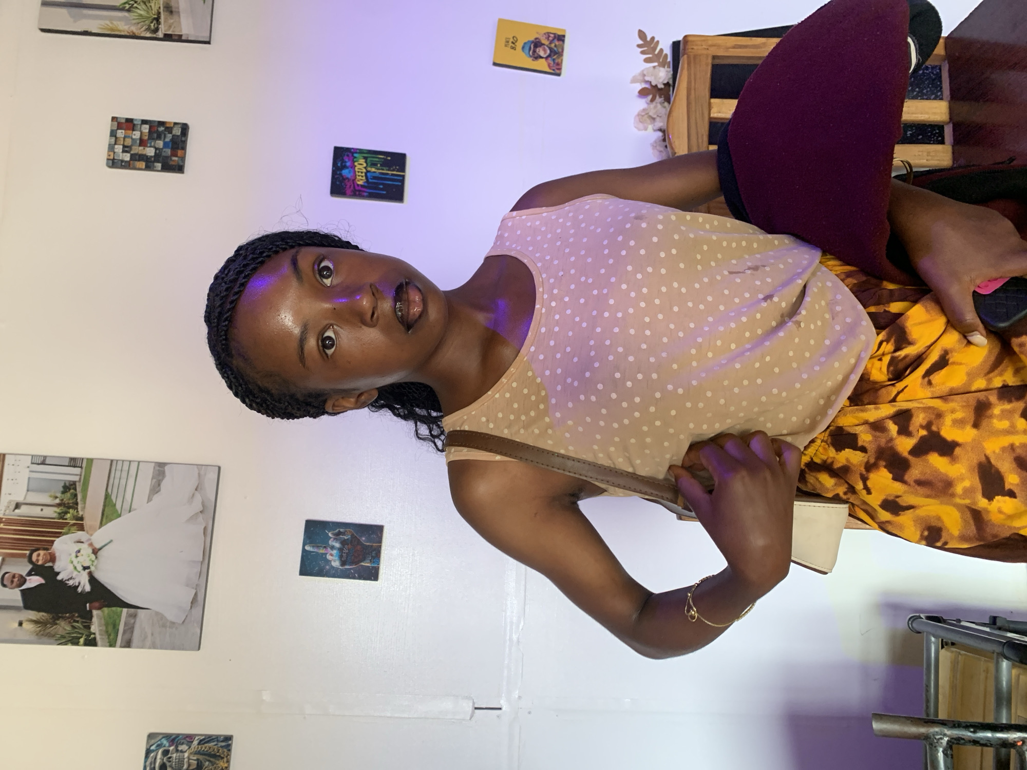 The image displays a young Black woman indoors, likely in Jos, Nigeria. She is positioned centrally, looking directly at the camera with a wide-eyed, slightly surprised expression and her mouth slightly open. She has dark skin and her hair is styled in braids.
She is dressed in a light beige or cream-colored tank top with small white polka dots, paired with a skirt featuring a vibrant yellow and brown abstract or animal print pattern. A brown strap, possibly from a shoulder bag, crosses her body. On her left wrist, she wears a delicate gold-colored bracelet. Her right hand appears to be holding a small white object, possibly a phone or a compact purse.
The background features a white wall adorned with several pictures and artworks. These include a picture of green foliage, a rectangular mosaic of colorful squares, and a black canvas with "FREEDOM" written in colorful, dripping letters. Above this, a small yellow object or picture reads "THAT BRO". To the left, a prominent wedding photograph shows a couple. Nearby, there's a picture of a colorful fist gesture and another with a skull design.
To the woman's right, a wooden chair or stool with a dark reddish-brown cushion is partially visible. Parts of a metallic furniture frame are seen in the bottom right corner. The lighting in the room has a distinct purple or blue hue, suggesting artificial light that creates a specific ambiance. The woman is posing for the photograph.
