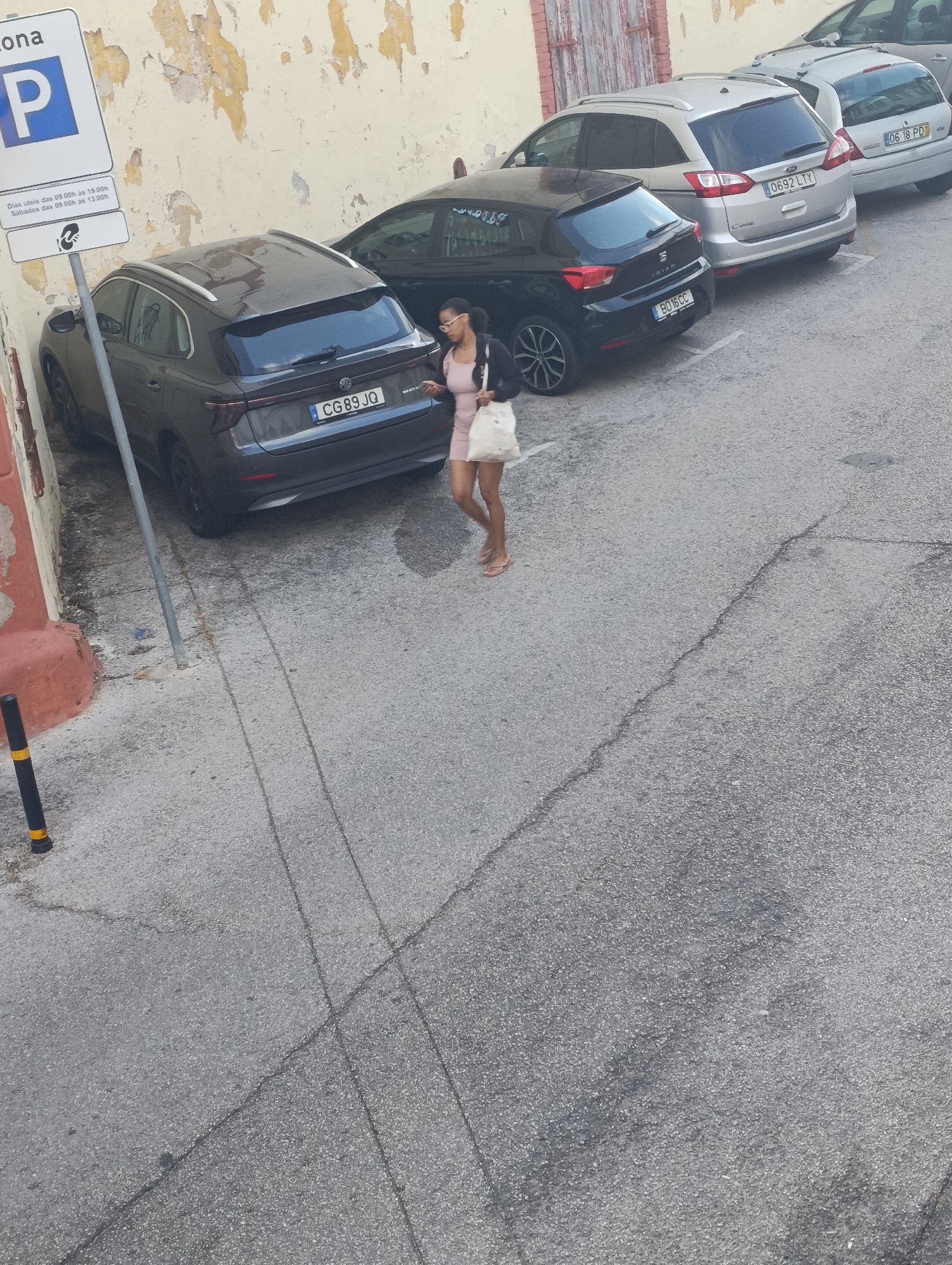 In Setúbal, Portugal, a woman walks past parked cars on a paved street next to a building with peeling paint. She is wearing a pink dress and carrying a white bag. The license plate on the gray car in the foreground reads "CG 89 JQ". A parking sign is visible on the left side of the image.