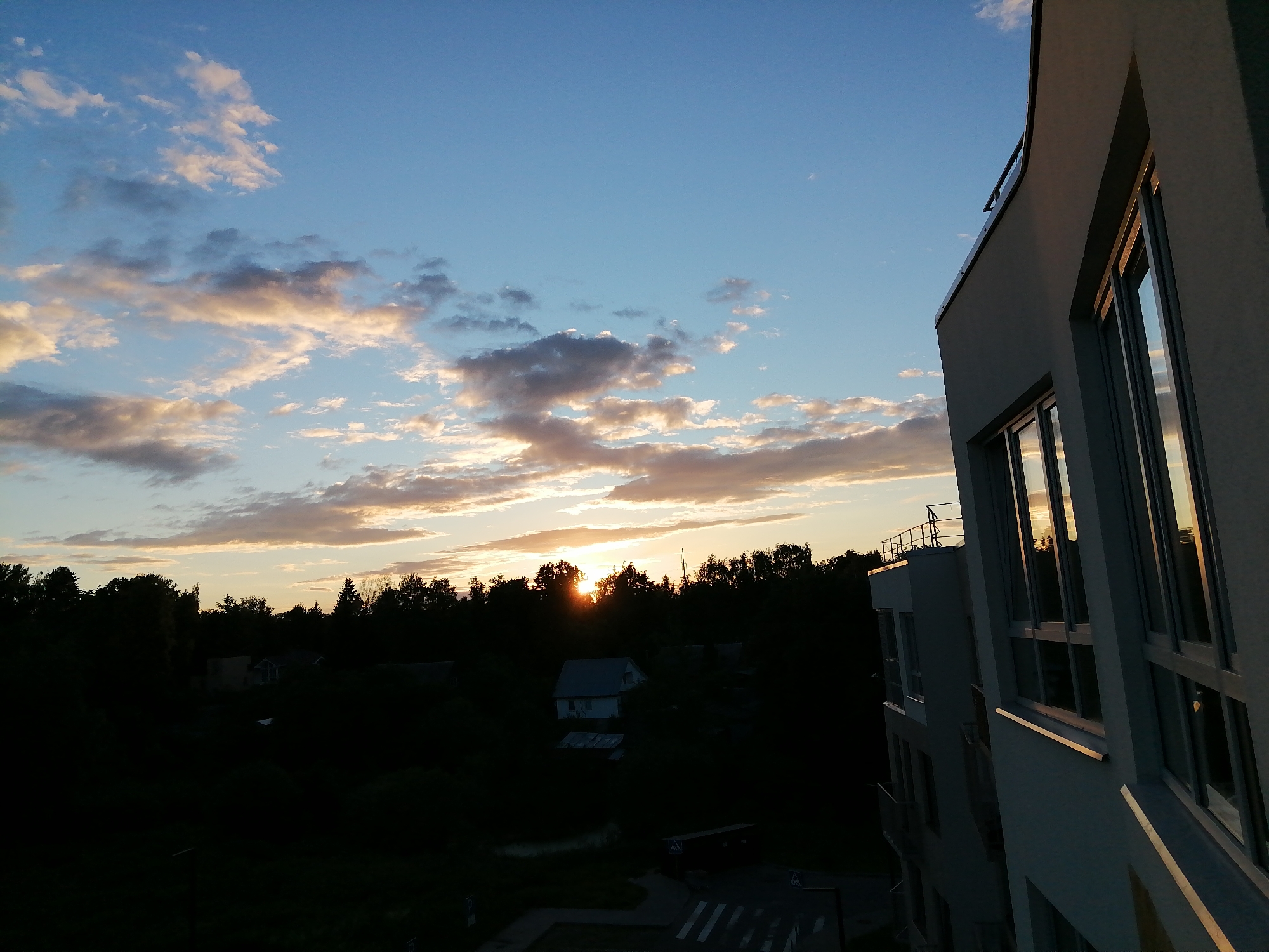 The image captures a sunset over a residential area. A modern white building with large windows dominates the right side of the frame, reflecting the warm hues of the setting sun. In the background, a dense line of trees separates the buildings from the sky, with the sun partially visible behind them, creating a silhouette effect. A few houses and smaller structures are visible nestled amongst the trees, partially obscured by darkness. The sky is a clear blue, dotted with scattered clouds that are tinged with orange and pink from the sunset. The overall scene suggests an urban or suburban landscape at dusk.