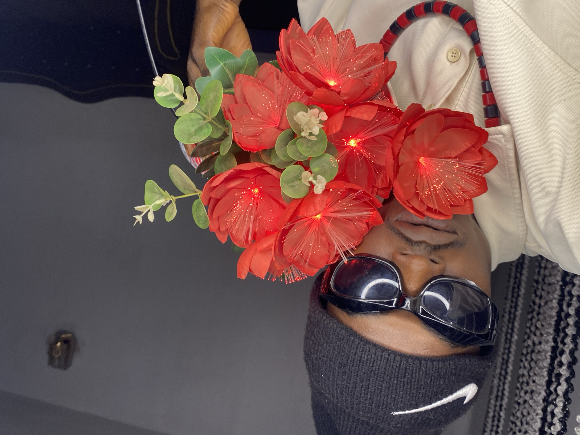 A young Black male, appearing to be in his late teens or early twenties, is depicted in a close-up, self-taken photograph. He is wearing a black Nike beanie, dark-lensed sunglasses with a thick frame, and a light-colored button-up shirt. A beaded necklace with alternating red and black cylindrical beads is visible around his neck, resting on the shirt. He is holding a bouquet of bright red artificial flowers with a fiber-optic glow in his right hand, which partially obscures his face. The background is a plain, dark gray wall, with a light fixture visible on the upper right side. The scene appears to be indoors, likely within a residential setting. There is no indication of the specific location in Abakaliki, Nigeria, beyond the inferred residential context.