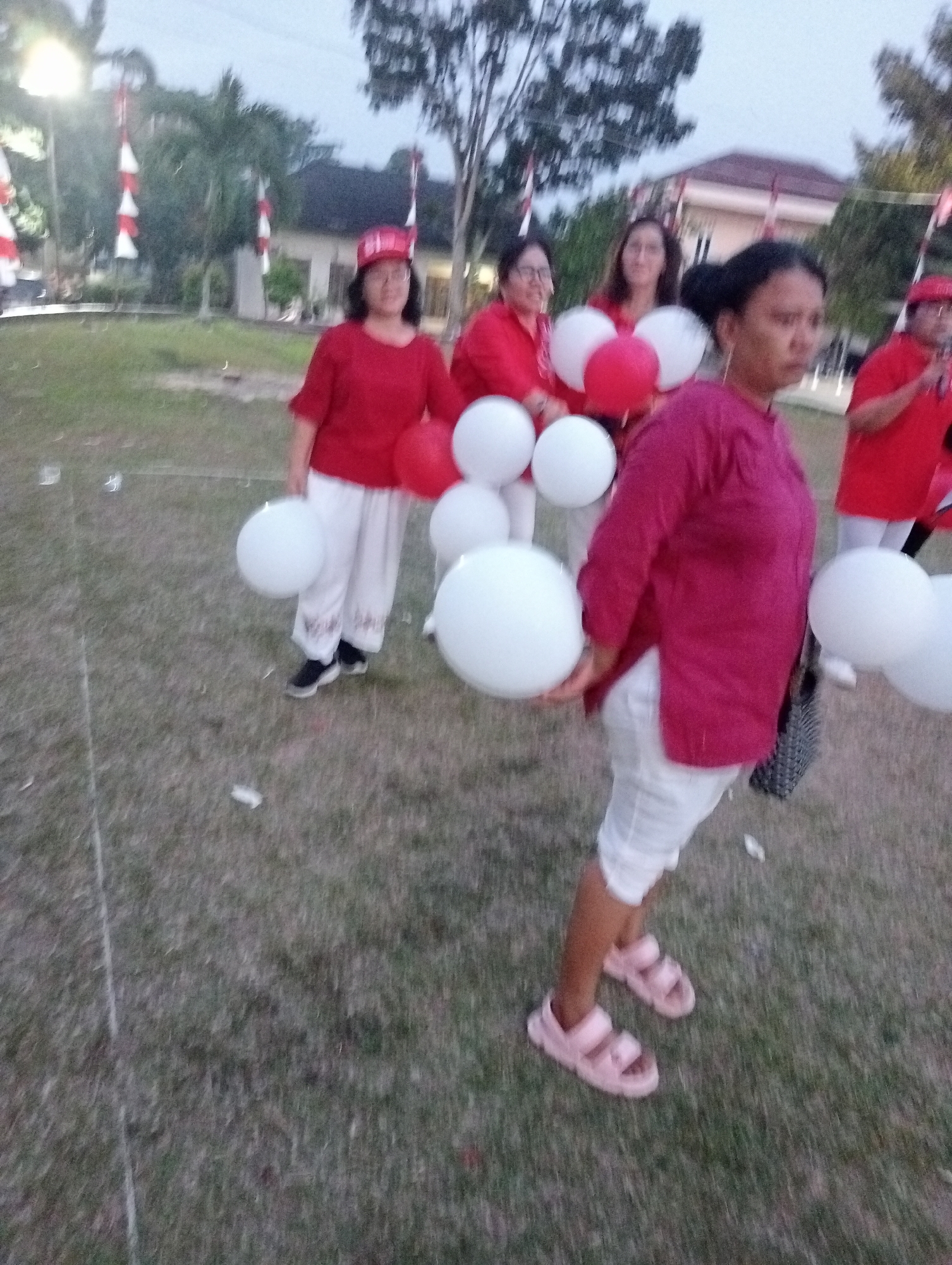 A group of women, some wearing red hats, are holding red and white balloons in an outdoor grassy area. The women are dressed in red shirts and white pants or skirts. In the background, there are trees and buildings, and Indonesian flags are visible hanging from poles. The lighting suggests it is dusk or early morning. The location appears to be a park or public space in Jambi, Indonesia, possibly during a celebratory event given the balloons and flags.