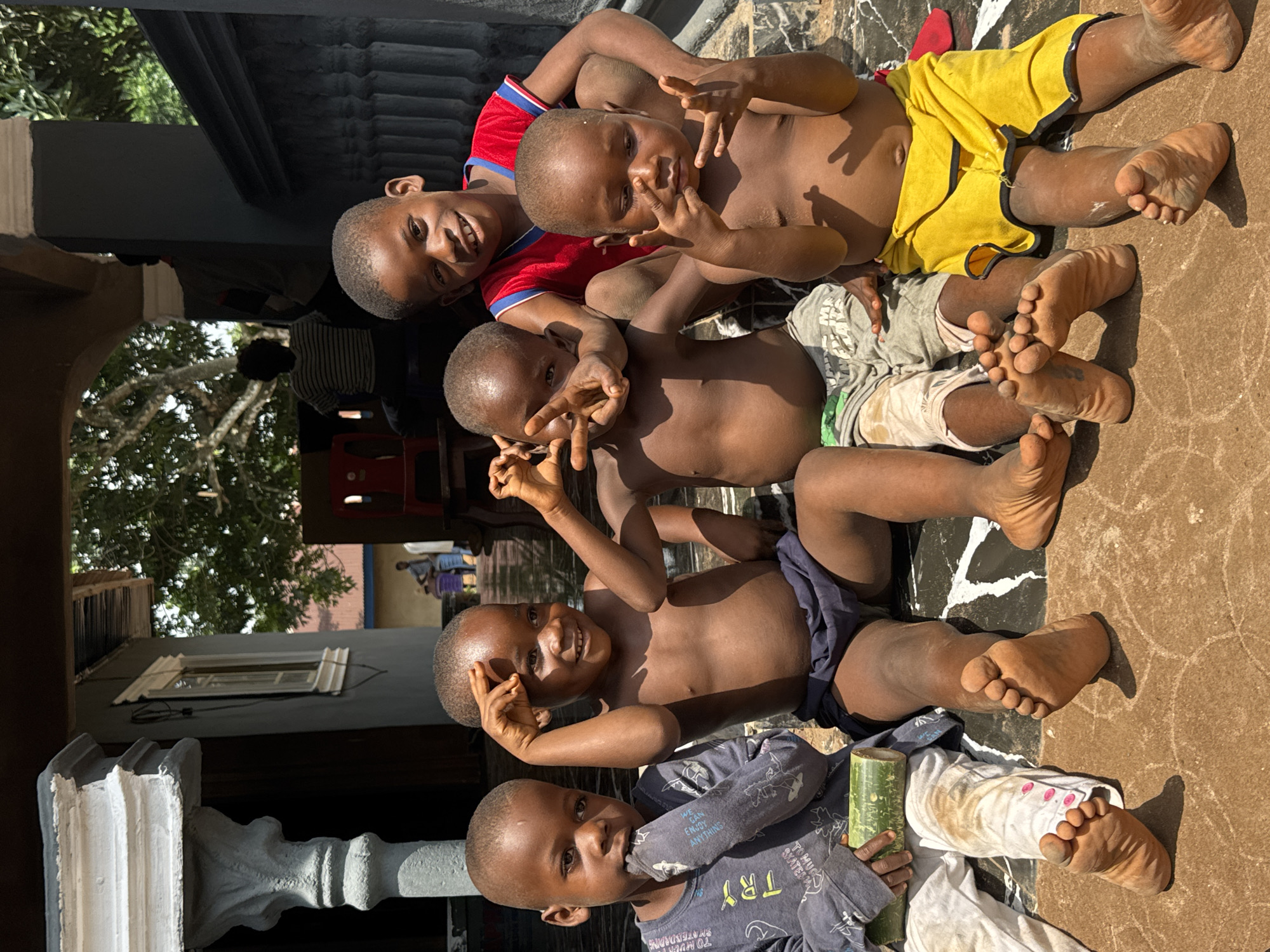 In Eziudo, Nigeria, a group of four young, dark-skinned children are lying down in a vertical stack on a brown, cracked surface, likely a patio or walkway. The top child wears a red shirt with blue trim and yellow shorts. The second child wears light green and grey shorts. The third wears dark blue shorts, and the bottom child is in a grey long-sleeved shirt and white pants. The surface they are on has a black and white painted design. The first three children are making hand gestures, the top two doing peace signs. The bottom child is holding a green cylindrical object. Behind them are elements of a building structure. A pillar is visible to the left, while elements of red chairs and a window are further back. There is tree foliage visible in the background.