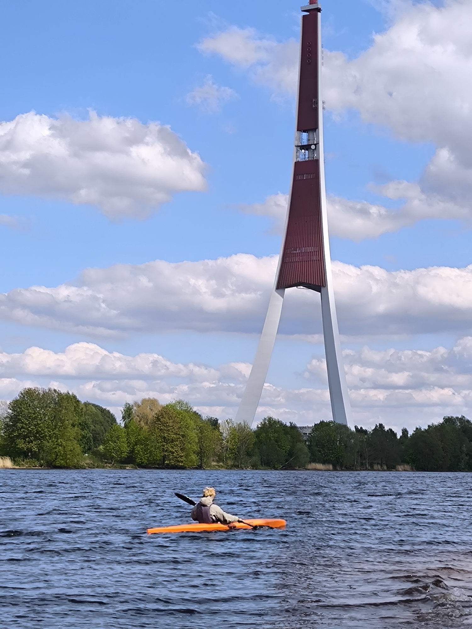 A medium, full outdoor shot captures a woman in a bright orange kayak on a choppy body of water, with the Riga Radio and TV Tower dominating the background. The woman is seen from the back, paddling with a black paddle. The tower is red and white, tall and slender, and is located in Riga, Latvia. A green tree line is situated on the water bank behind the kayaker. The sky is light blue, filled with fluffy, white clouds.