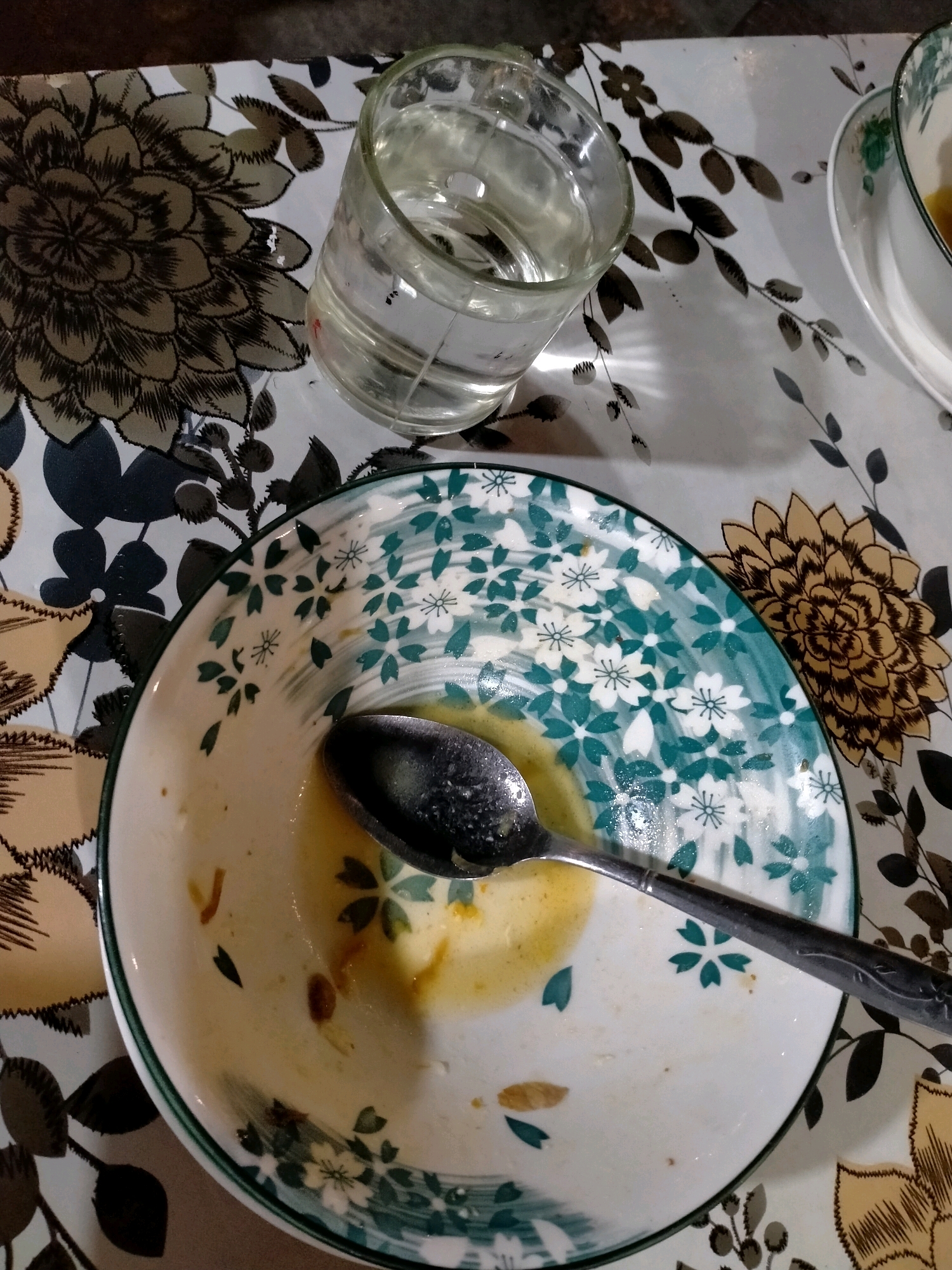 The image shows a close-up, overhead view of a partially eaten meal on a floral-patterned tablecloth. A used ceramic bowl with green and white floral designs contains remnants of yellow food and a spoon. To the left of the bowl, a clear glass filled with water sits on the tablecloth. The lighting suggests an indoor setting. The scene is devoid of any people or animals. The location of Ambon, Indonesia is not depicted or suggested by any elements in the image.