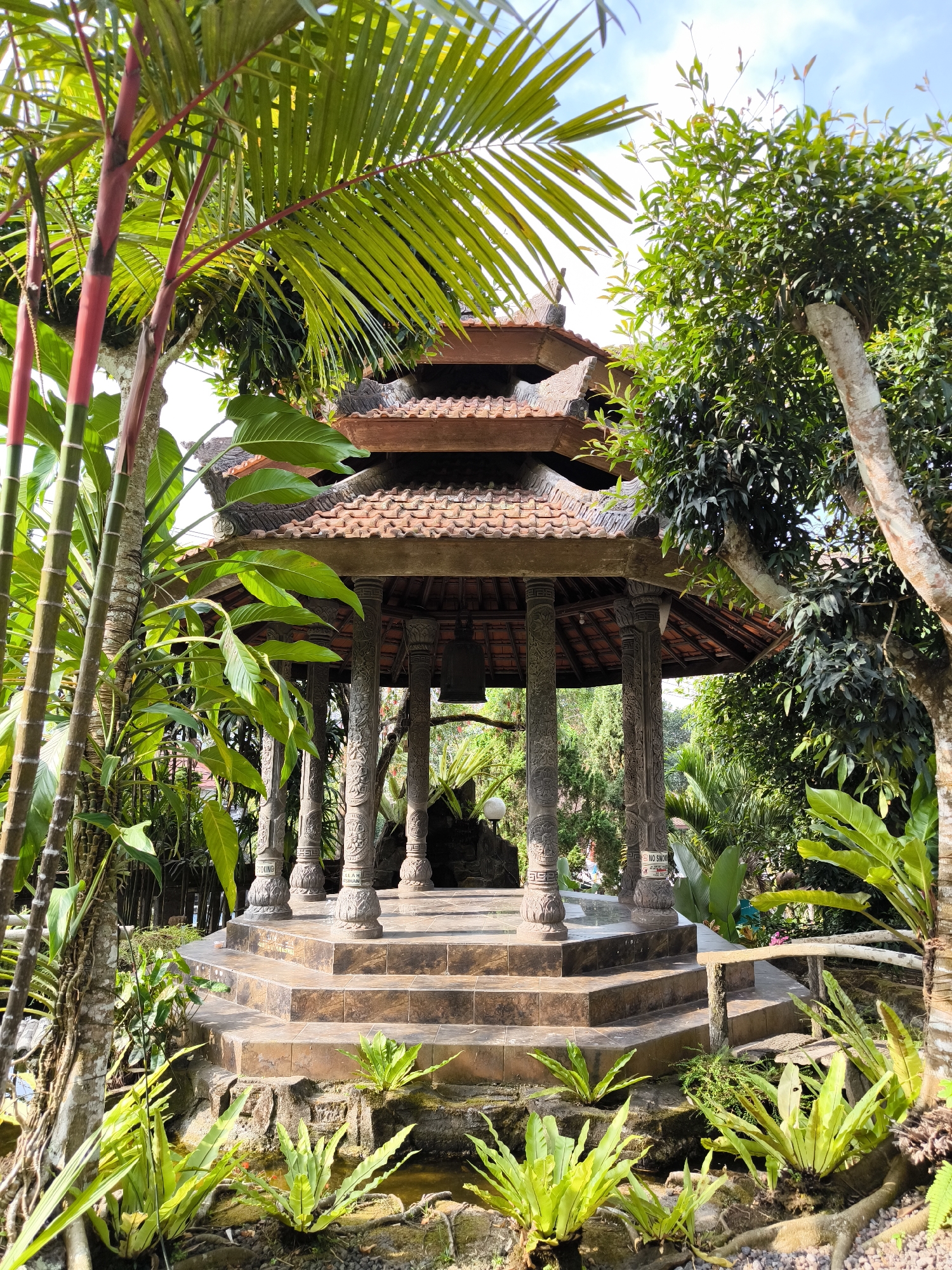 The image depicts a Balinese-style pavilion situated within a lush, tropical garden in Pupuan, Indonesia. The pavilion features a tiered, terracotta-tiled roof and is supported by elaborately carved stone pillars. The structure is elevated by a series of stone steps. Surrounding the pavilion are abundant green plants, including palm trees with striking red stems, ferns, and large-leafed foliage. The ground around the pavilion is a mix of stone, gravel, and shallow water features, with ferns growing along the edges. The scene is bathed in natural light, suggesting it is daytime. The image captures the tranquility and architectural beauty of the location.