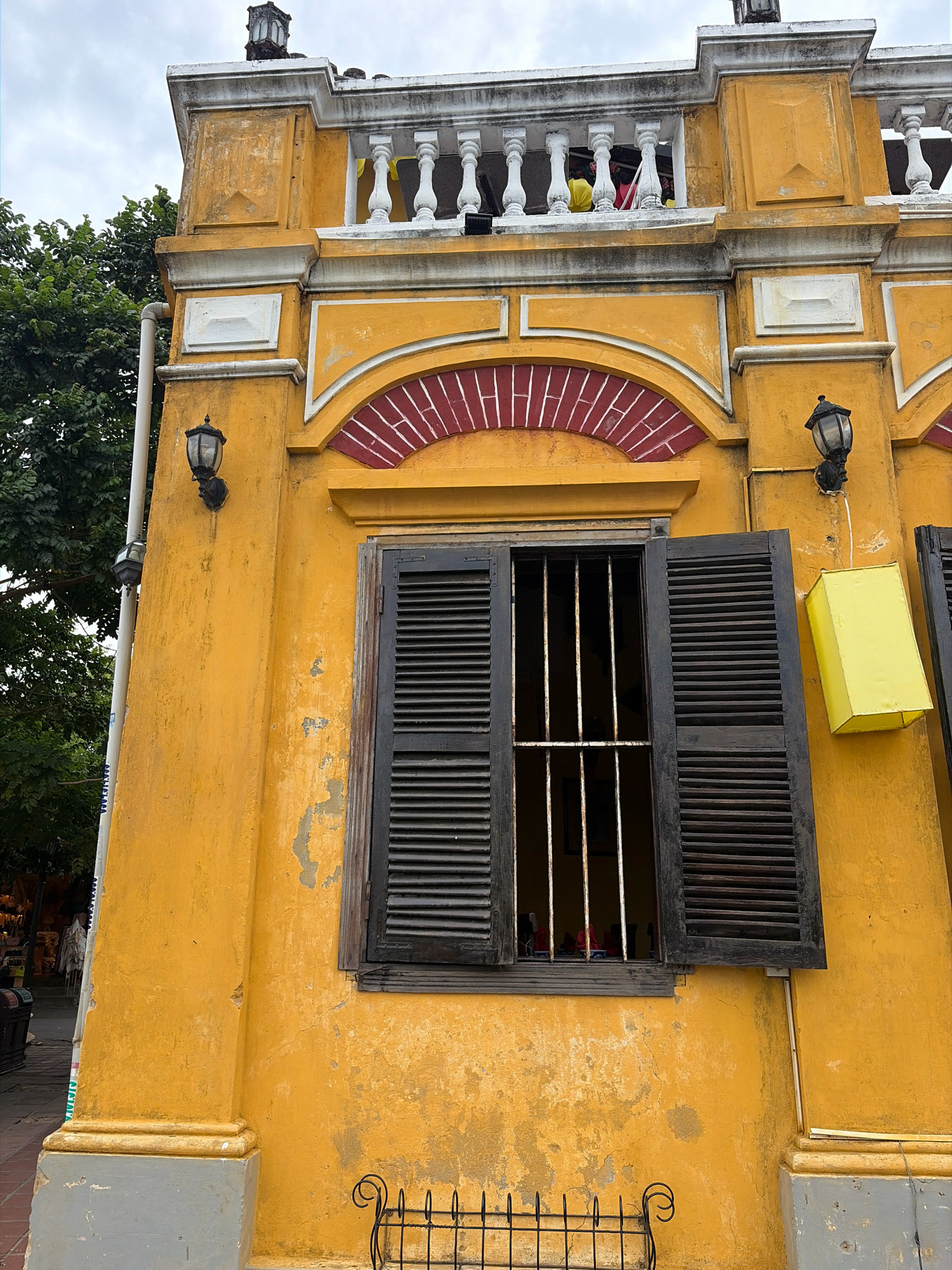 The image shows a yellow building with architectural details in Hoi An, Vietnam. The building has a weathered facade with peeling paint. A large wooden window with dark shutters is open, revealing metal bars. Above the window, there is a red brick arch. A bright yellow rectangular lantern hangs on the right side of the building. A white balcony with balustrades is visible at the top of the building, and people can be seen on the balcony. The sky is overcast. Green foliage from trees is visible on the left side of the image. The lower part of the building has a gray base.