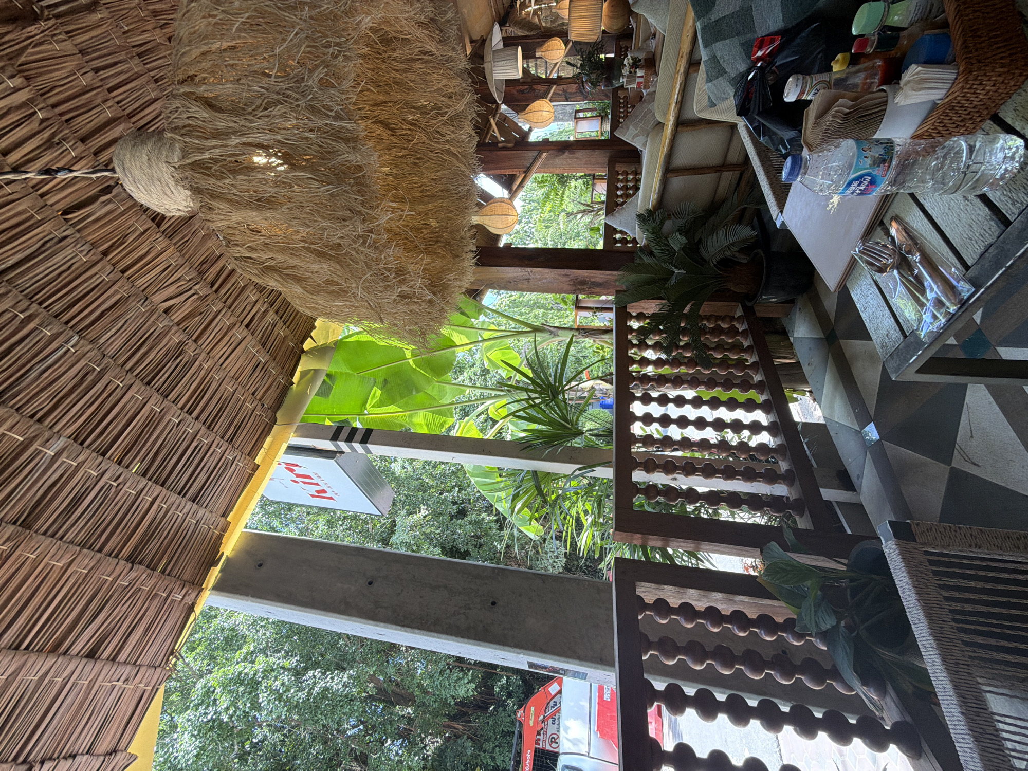The image depicts an outdoor seating area of a restaurant or cafe, likely in Karon, Thailand, with a thatched roof overhead and lush greenery visible in the background. A large, natural fiber lampshade hangs from the ceiling. In the foreground, a wooden table is set with cutlery in plastic packaging, a bottle of water, napkins, and various condiments including ketchup and hot sauce in a basket. A patterned tiled floor is partially visible. A wooden railing with decorative spindles separates the seating area from a walkway. Beyond the railing, large tropical plants and trees are abundant. A red vehicle and construction equipment are visible in the far background, suggesting proximity to a street. The overall ambiance is relaxed and tropical.