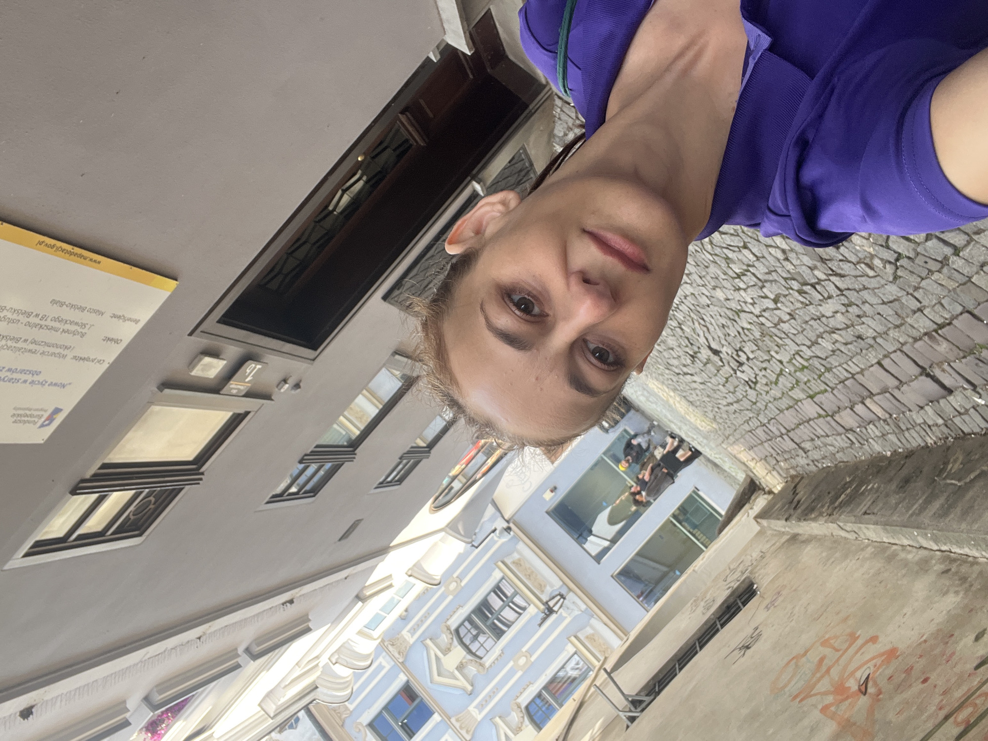 An outdoor, eye-level, medium shot captures a woman in a purple collared shirt taking a selfie. She is facing the camera, angled slightly to the right. The woman has light skin, brown eyes, and dark hair pulled back. Her right ear is visible. Her mouth is slightly parted, and she is smiling.
The background depicts a narrow cobblestone street lined with buildings. The architecture on the left side of the street is light blue with white decorative accents and multiple windows. On the right, a gray building features dark-framed windows and a dark wooden door. A sign with text and a website address is affixed to the gray building. The street itself is paved with gray cobblestones. In the distance, on the left, a few people are visible walking down the street. The lighting suggests it is daytime.