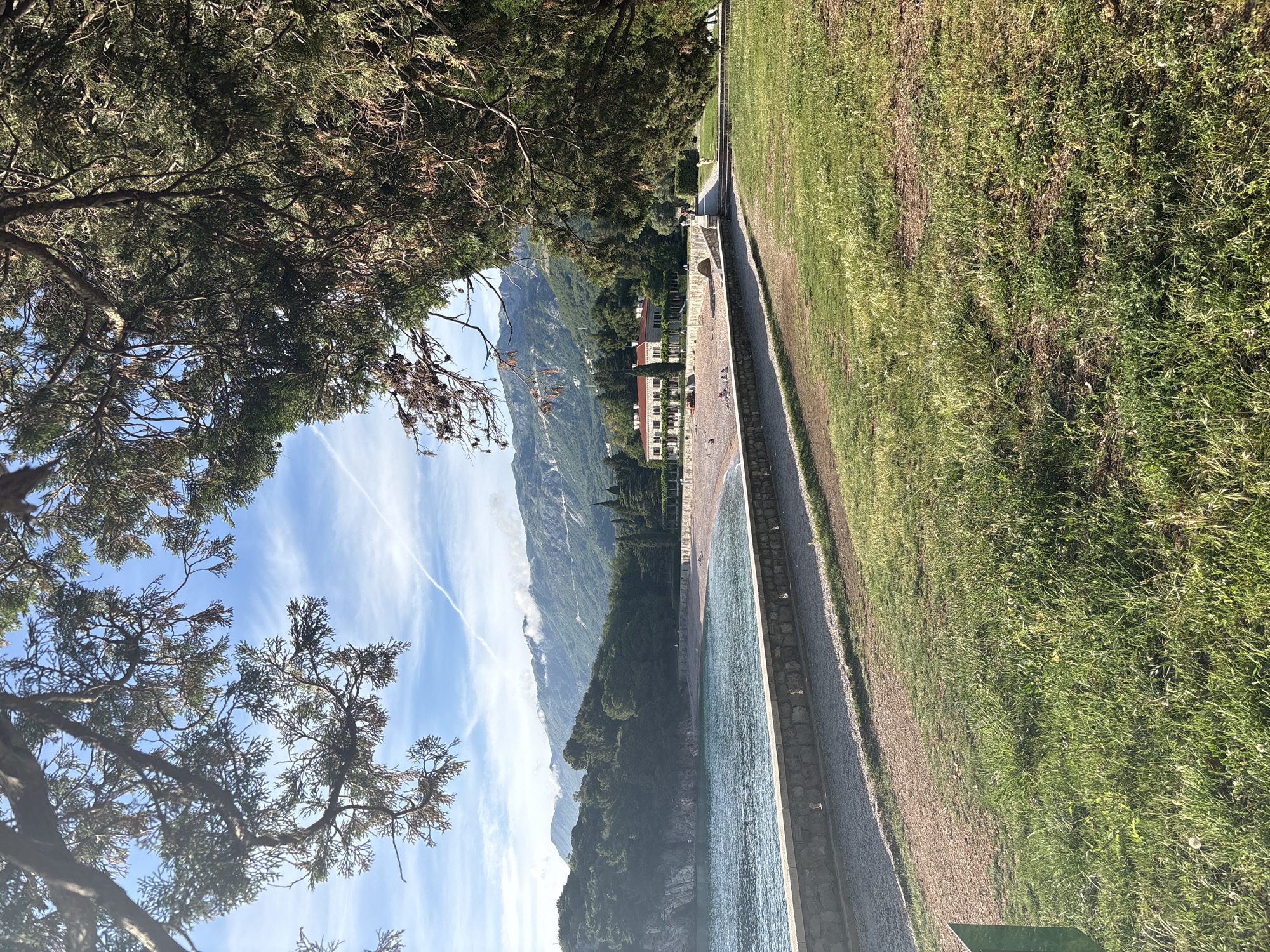 A scenic view in Sveti Stefan, Montenegro, shot from a ground level perspective, is visible through tree branches in the foreground. The blue sky with wispy clouds is visible through the branches. In the middle ground, the beach line runs alongside a long building with a red roof. Mountains rise in the background, partially covered in trees. To the right of the beach, there is a grassy lawn and a stone path.