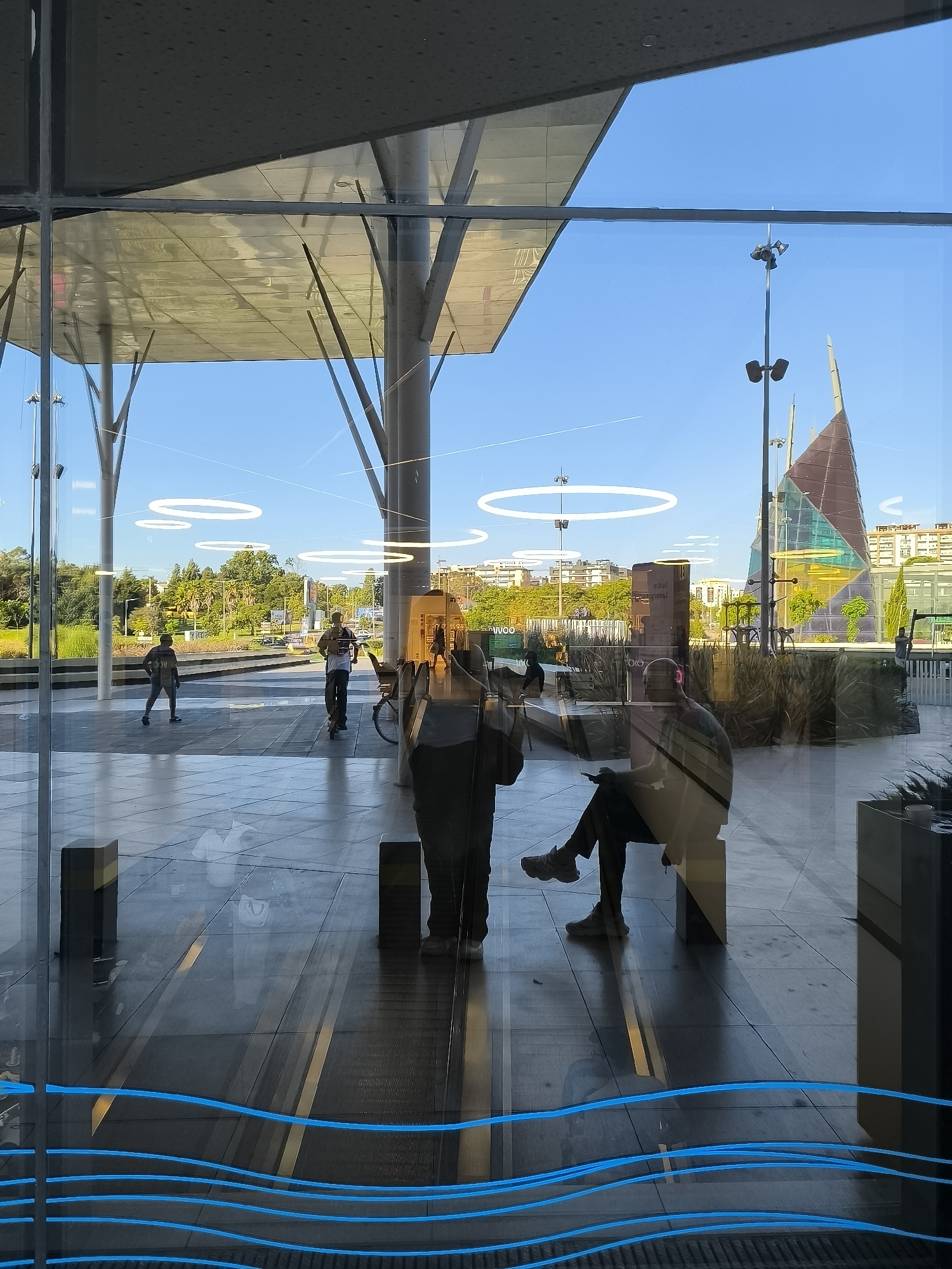 This image captures a reflection on a glass surface in Setúbal, Portugal, likely at a modern public transportation hub or a plaza. The reflection shows a bright, sunny day with a clear blue sky. In the foreground, the glass has a decorative blue wave pattern.
Reflected within the glass, we see the exterior architecture, characterized by a large, angled, opaque roof supported by slender, modern columns. Beyond this structure, the sky is visible, with several prominent circular light fixtures suspended in the air. A tall lamppost with multiple lights stands on the right side of the reflection, and further in the background, a distinctive, sail-like modern sculpture with colored glass panels is visible. The cityscape behind it consists of buildings and trees.
Within the scene reflected, several people are present. On the left, a person is riding a scooter. Slightly to the right of center, another person is also riding a scooter. Towards the right side, two individuals are seated, one of whom appears to be looking at a phone. The ground is paved with large tiles, and there are some planters with greenery. The overall impression is one of a contemporary urban space, bathed in sunlight.