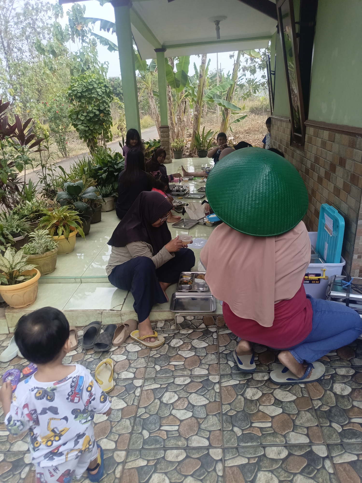 A group of people are gathered outdoors, possibly in the city of Karangkedawung, Indonesia, to eat a meal. Several individuals are seated on the tiled floor, while others are on a raised platform. A young child in a white shirt with construction vehicle prints stands in the foreground, looking at the food. In the mid-ground, a person wearing a large green conical hat and a pink headscarf is seated on the floor, facing away from the camera. A person in a black hijab is also seated on the floor to the left, holding a utensil. Other individuals, including children, are visible in the background, some seated and some standing. Various food containers and plates are arranged on the floor and a metal tray. The environment appears to be a covered outdoor area, likely a porch or veranda, with a tiled floor and a green-painted ceiling. Potted plants are situated along the edges of the platform, and vegetation, including banana trees, is visible in the background. A light-colored wall with a patterned tile lower section is present on the right side of the image.
