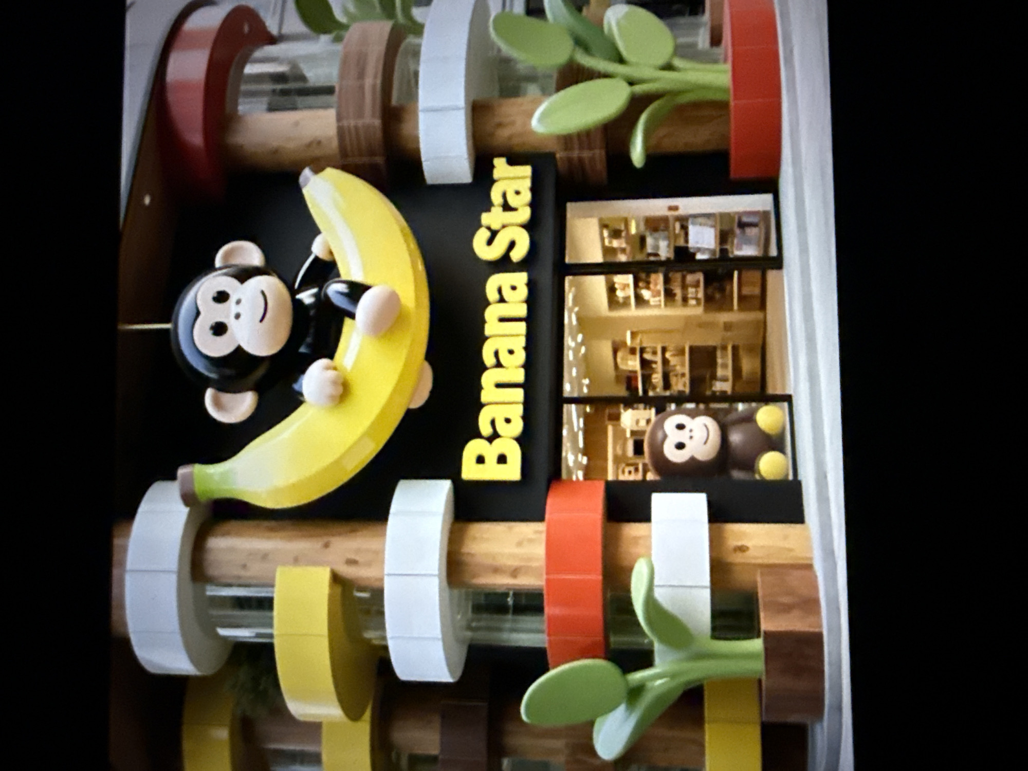 The image depicts the exterior of a store named "Banana Star" in Taipei, Taiwan. The store features a playful design with a large, yellow banana sculpture as a prominent element of its facade. A cartoon-like monkey character is seated on the banana, appearing to be part of the store's branding. The store's name is displayed in bright yellow lettering on a black background above the entrance. The entrance itself appears to be a glass door leading into a well-lit interior with shelves filled with merchandise. Surrounding the entrance are decorative columns with colored bands of white, yellow, and red, and wooden accents. On either side of the entrance, there are stylized green plant sculptures in wooden and red planters. A small, brown monkey figure is positioned to the left of the entrance, near the plant sculpture. The overall aesthetic suggests a retail establishment targeting a younger demographic or those interested in whimsical themes. The background shows more of the building's structure with visible windows and architectural details.
