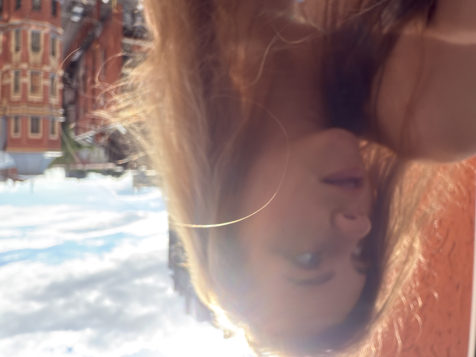 The image is an upside-down, close-up selfie of a young woman with blonde hair. She is outdoors, and the background suggests an urban setting with buildings and a cloudy sky. The woman's face is partially obscured by her hair and the angle of the shot, but her eyes, nose, and lips are visible. She appears to be looking towards the camera, and her expression is neutral. A thin, circular earring is visible in her left ear. The lighting suggests it is daytime, possibly late afternoon due to the warm tones. The buildings in the background are ornate and appear to be made of brick, with multiple windows. The sky is filled with soft, white clouds. The overall mood is casual and personal. There is no visible text in the image.