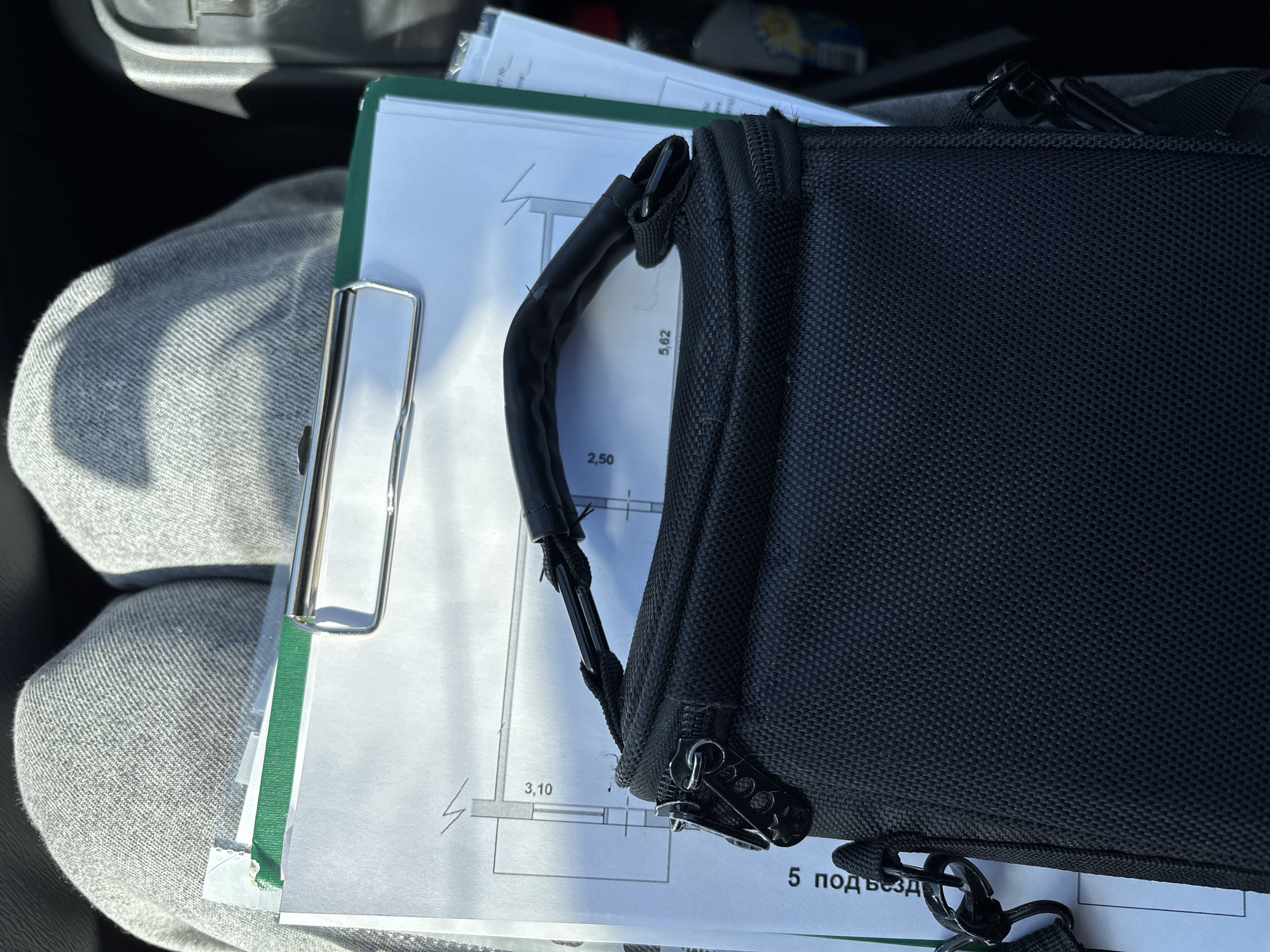 The image captures a close-up, high-angle view of a person's lap, covered by grey, textured fabric trousers, likely sweatpants or joggers. Resting on the lap is a green-backed clipboard holding several white papers, secured by a prominent silver metal clip. Overlapping these documents is a black, woven fabric bag, possibly a small utility case or laptop bag, featuring a padded handle and two prominent zippers with round pull tabs.
The visible documents display technical or architectural drawings, including lines, numerical dimensions such as "2,50", "3,10", and "5,62", and a lightning bolt-like symbol. Partially legible Cyrillic text at the bottom right reads "5 подъезд," which translates to "5 entrance" or "5 stairwell." Bright natural sunlight illuminates the scene, suggesting it is daytime. The background is dark and out of focus, indicating the setting is likely inside a vehicle, with glimpses of a dark car interior visible in the upper left. The scene suggests someone reviewing work-related documents, possibly while in transit or waiting in Ust-Ilimsk, Russia.