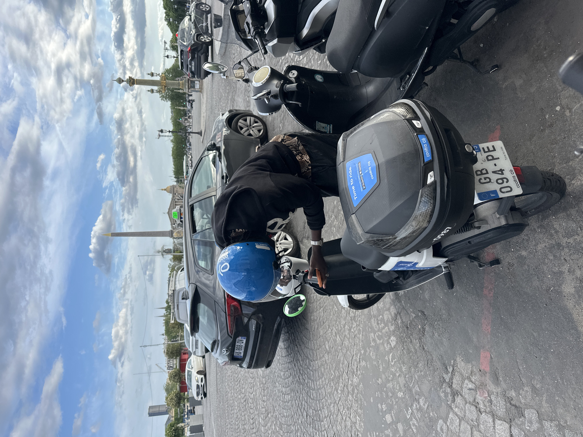 A ground-level outdoor shot shows a person wearing a blue helmet and a black hoodie interacting with an electric scooter parked on a cobblestone surface. Several vehicles, including cars and scooters, are parked nearby. In the background, the Luxor Obelisk stands tall in the Place de la Concorde, Paris, under a cloudy sky. The scooter has a license plate reading "GB-094-PE," and various "cooltra" logos are visible on the scooter and helmet.
