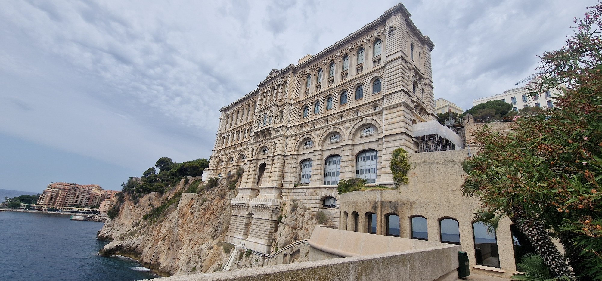 The image depicts a grand, ornate building, likely a museum or administrative structure, situated on a steep cliff overlooking the Mediterranean Sea in Monaco-Ville, Monaco. The building, constructed of light-colored stone, features multiple stories with numerous arched windows. Its architecture is classical, characterized by decorative molding and columns.
In the foreground, a grey concrete walkway and wall create a boundary, leading the viewer's eye towards the impressive edifice. To the right, lush green foliage and a palm tree with a distinctive patterned trunk partially obscure the view of the building's lower sections. The sea, a deep blue, is visible to the left, with a coastal town characterized by terracotta-colored buildings in the distance.
The sky is partly cloudy, with wispy white clouds against a pale blue backdrop, suggesting a moderate day. Scaffolding and construction materials are visible on the side of the main building, indicating ongoing renovations or expansions. The overall scene conveys a sense of historical significance and architectural grandeur, set against the dramatic backdrop of the Monegasque coastline.