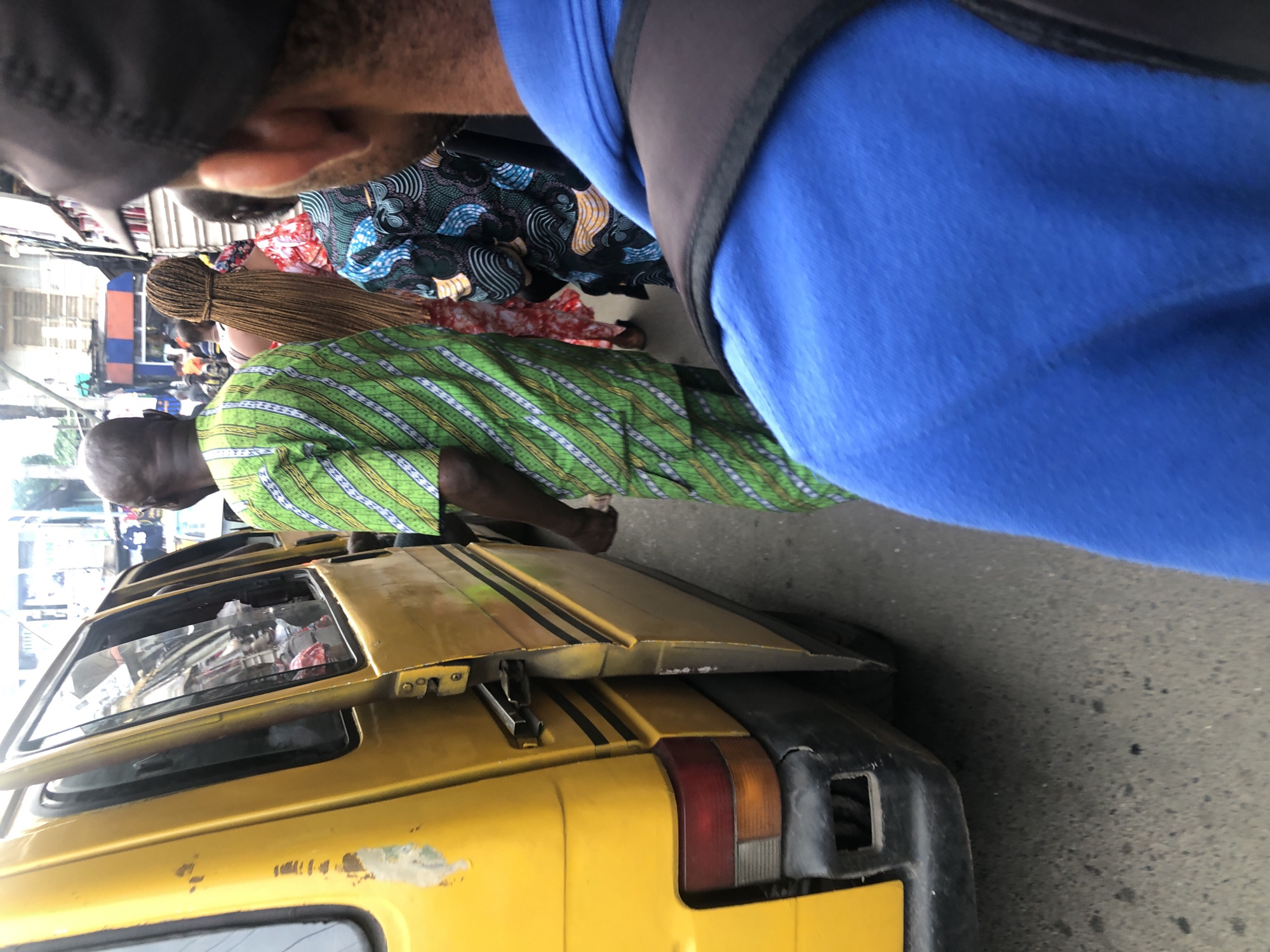 The image depicts a street scene in Ikeja, Nigeria, with a yellow van occupying the left side. A man in a green and white striped garment stands next to the van. Further back, a person with long, braided hair is visible, along with others in colorful attire. The environment appears to be an urban setting with buildings in the background. The perspective is from behind two individuals, one in a blue shirt and the other in a black cap. The ground is paved with asphalt.